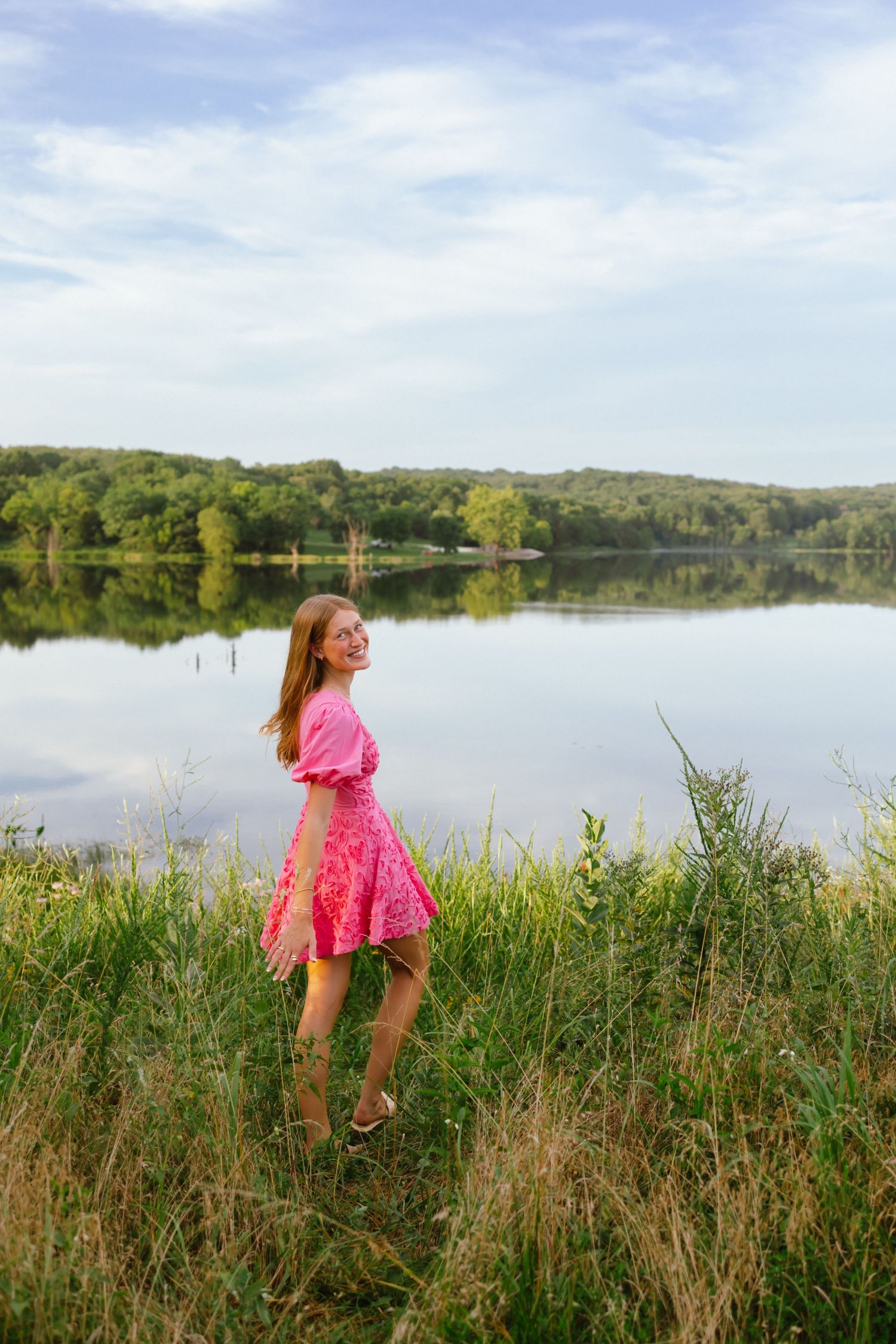 Young woman in pink dress standing by a calm reflective lake surrounded by lush green trees on a sunny day.