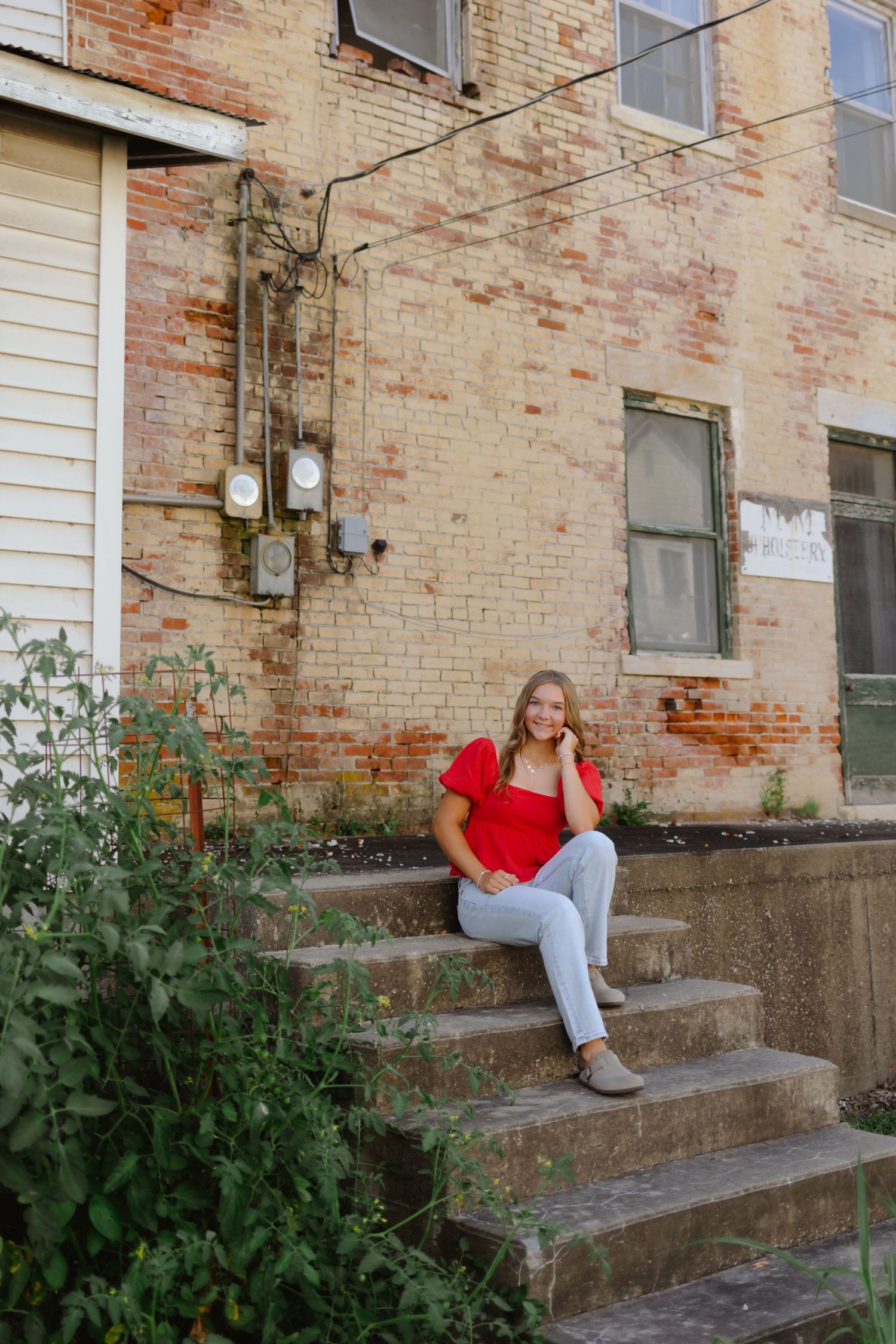 A person in a red top and light pants sits casually on concrete steps against a weathered brick wall.