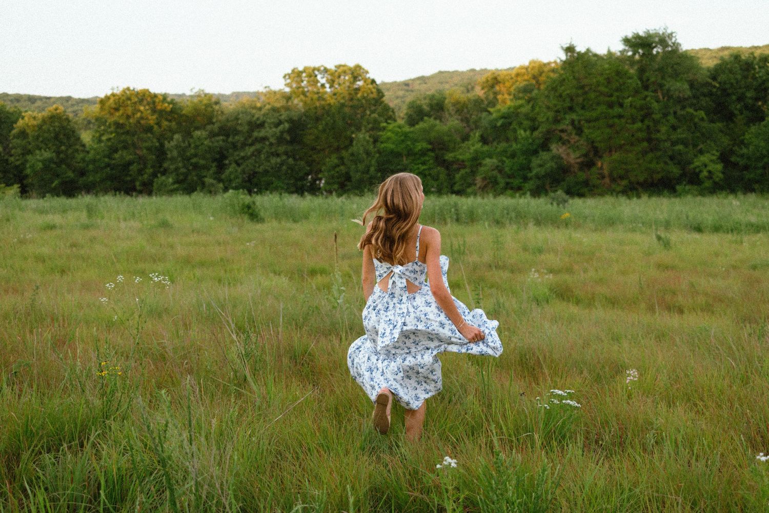A young girl in a floral dress runs through a lush green meadow with trees and hills in the background.