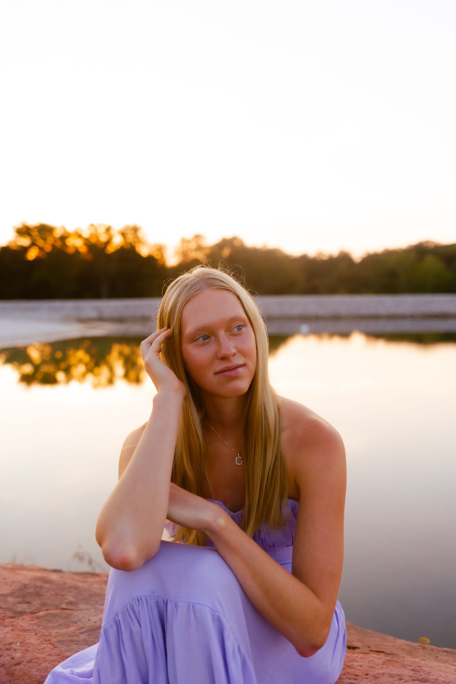 Young woman in purple dress poses by tranquil lake during golden sunset.
