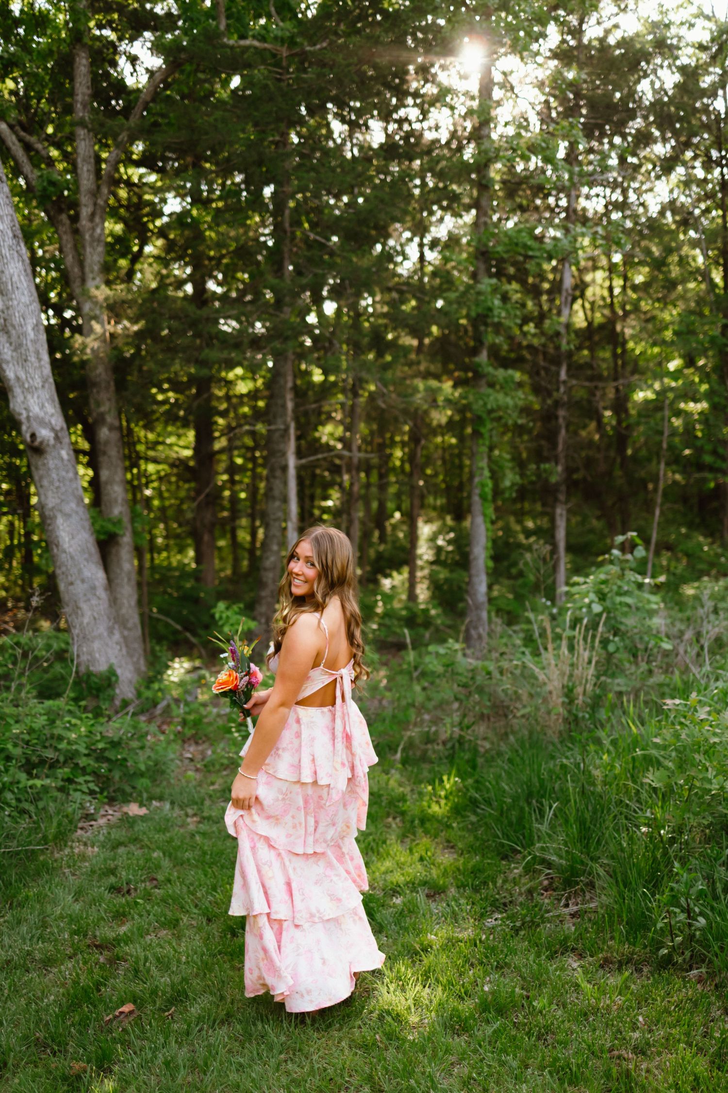 A young woman in a pink ruffled dress holds a colorful bouquet while standing in a lush green forest, smiling over her shoulder.