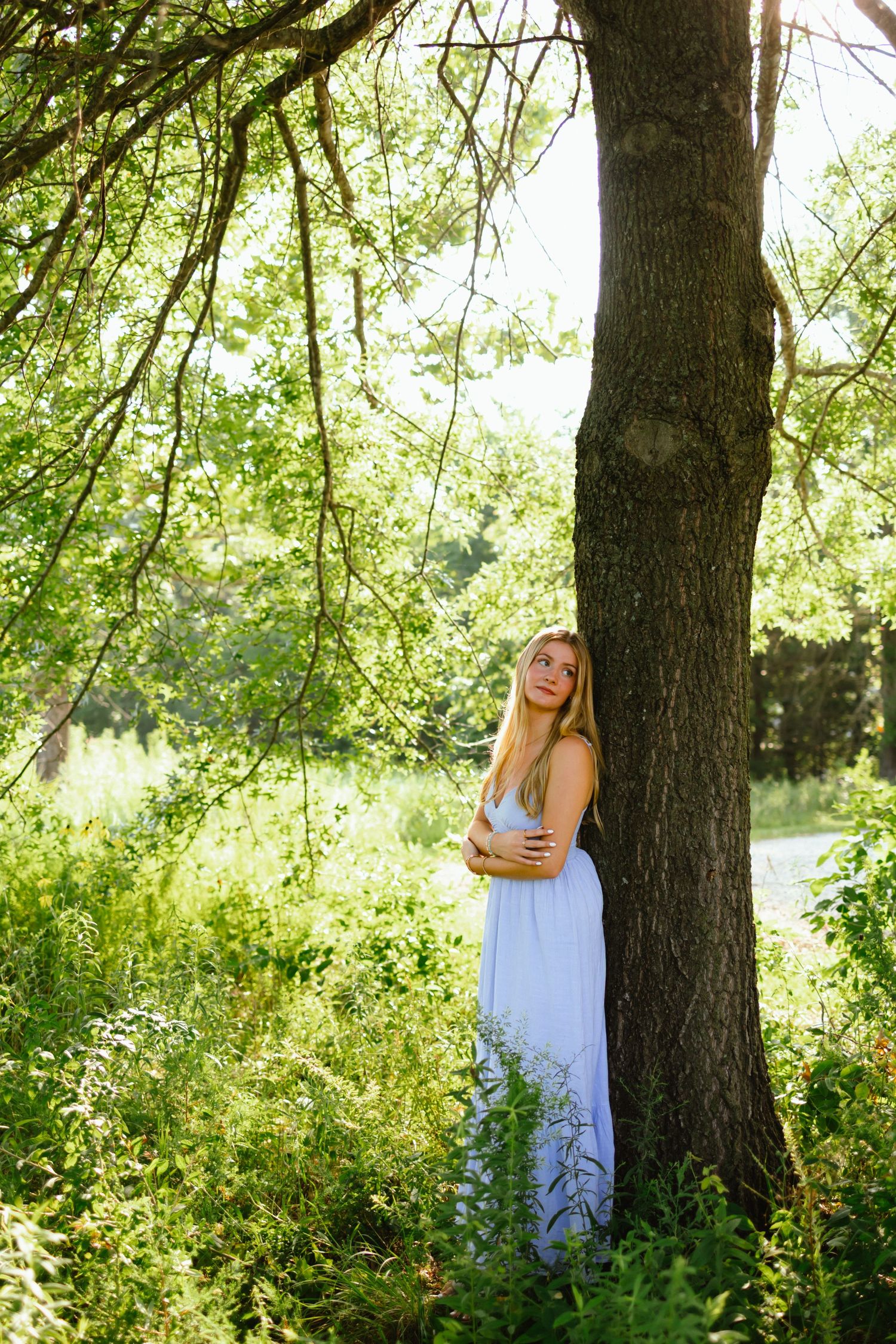 Young woman in white dress standing by large tree trunk in lush green forest with sunlight filtering through leaves.