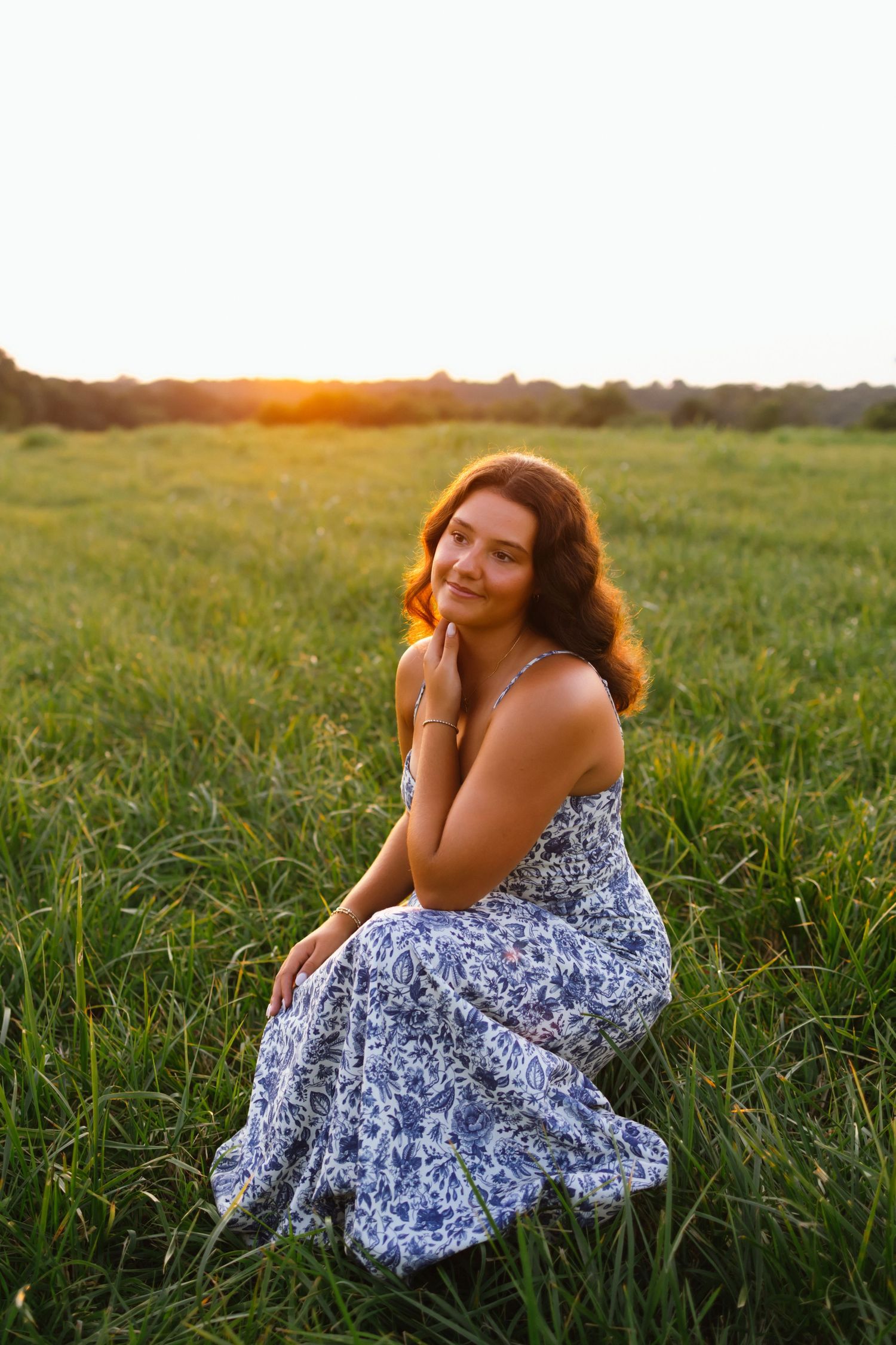 Young woman in floral sundress sitting in green field at golden sunset, smiling softly with hand on cheek.