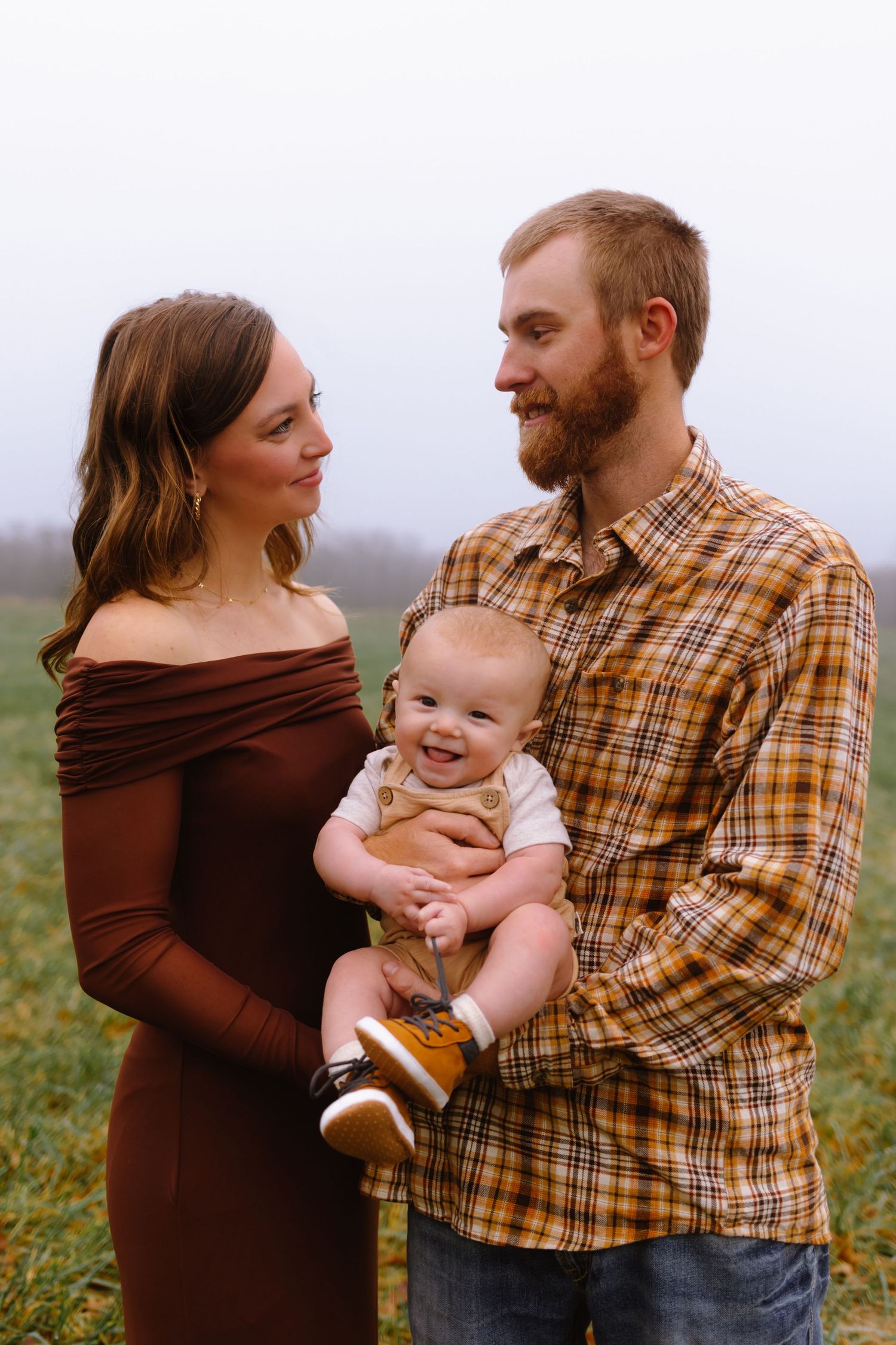 A couple in a brown sweater and plaid shirt hold their baby while standing in a grassy field at sunset.