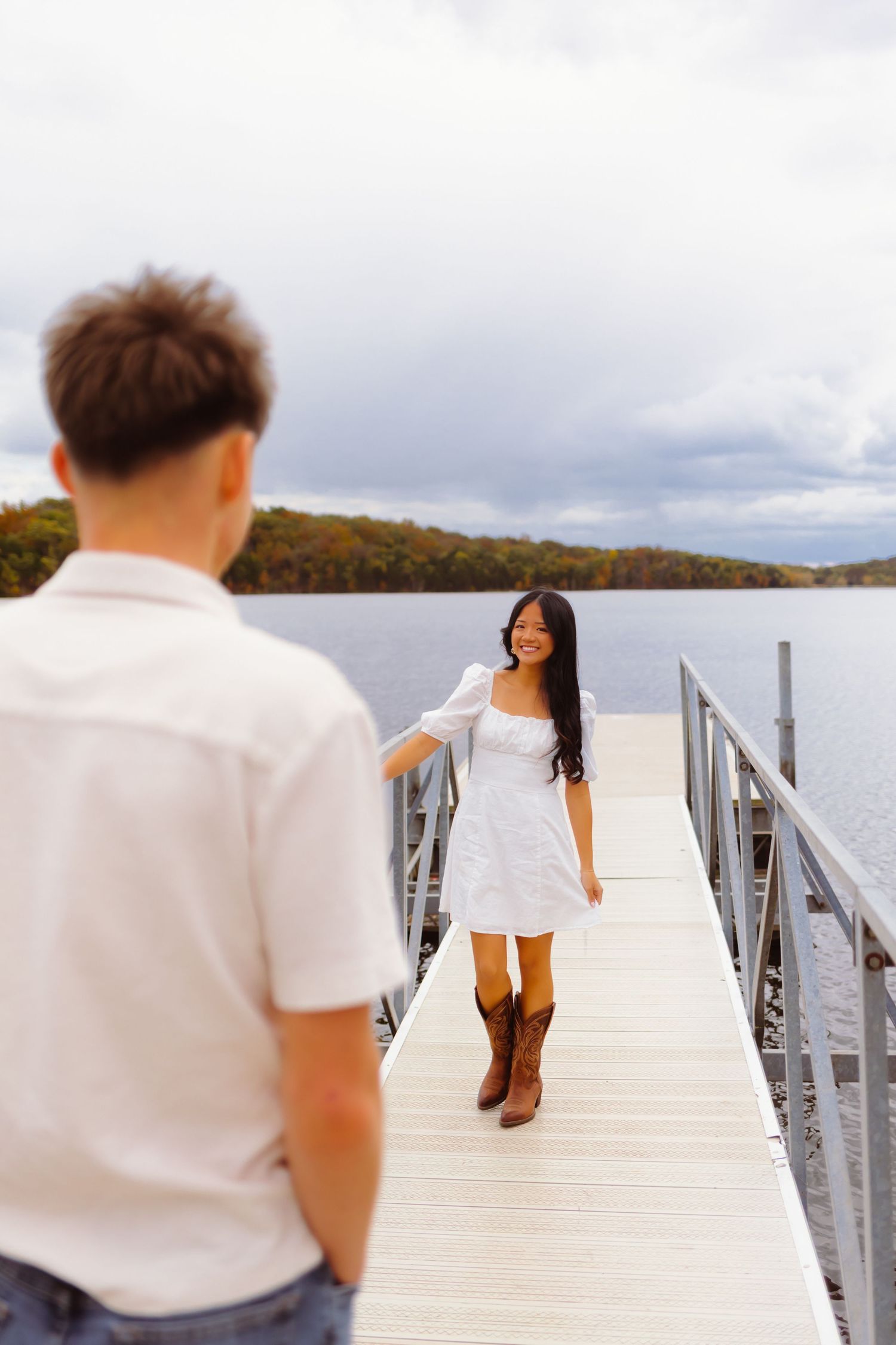 A couple stands on a wooden pier overlooking a serene lake with cloudy skies in the background.
