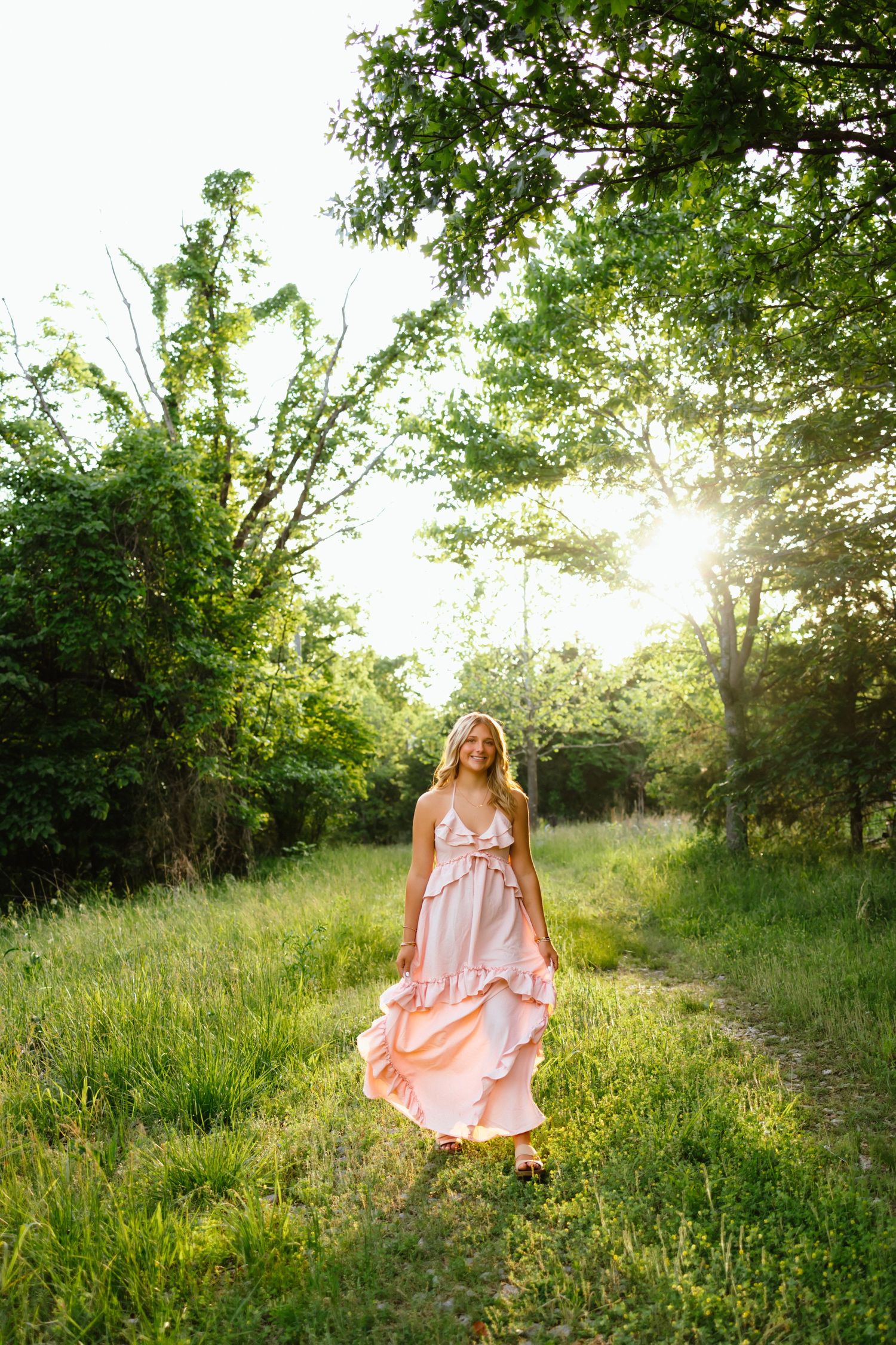 Young blonde woman in a flowing pink ruffle maxi dress walking through a sunlit green forest path.