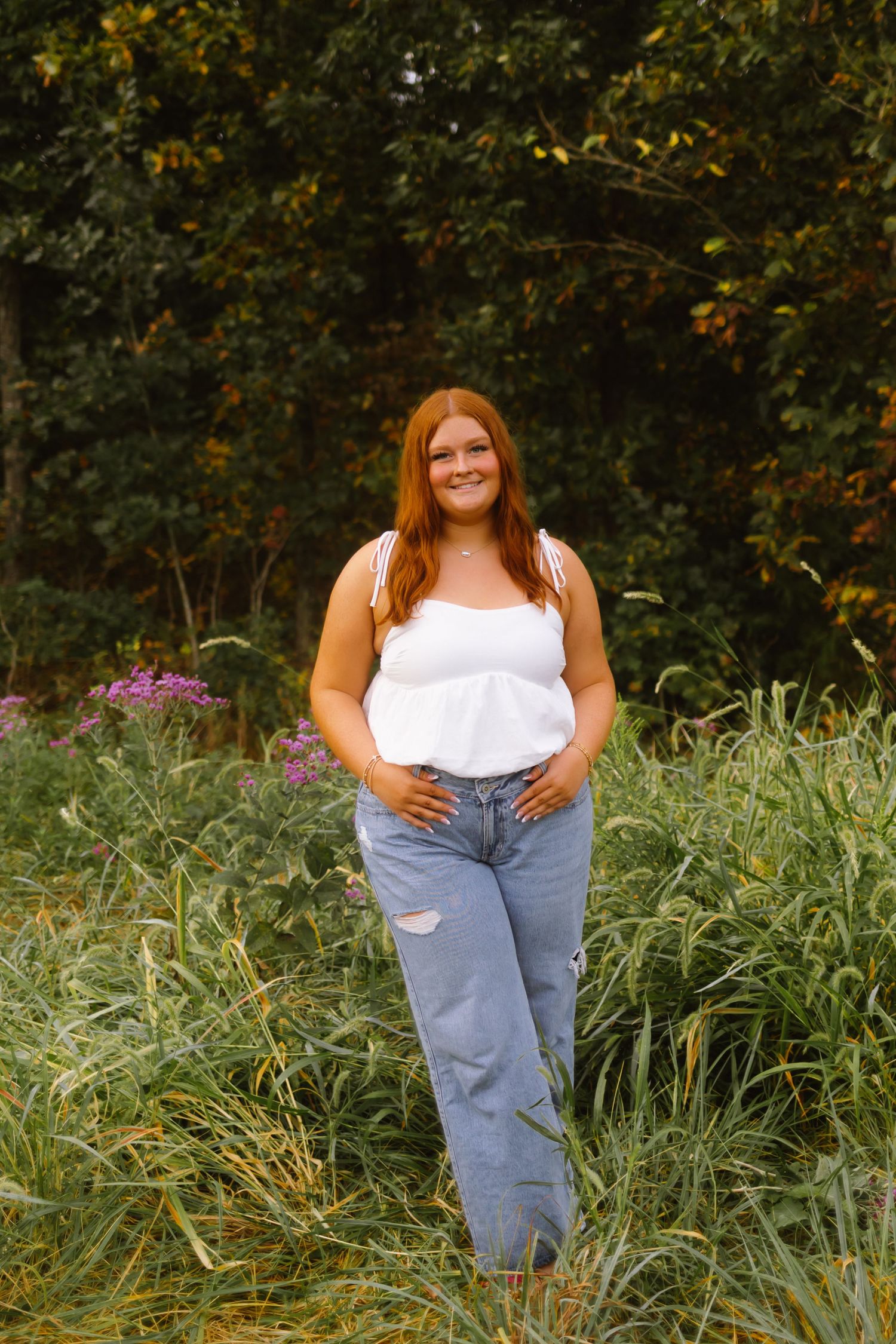A person in a white top and distressed jeans poses outdoors among lush greenery and flowering plants.