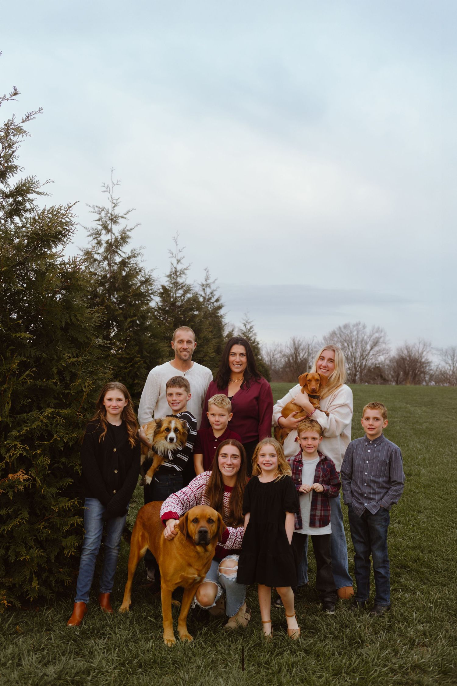 A group of people and a large brown dog pose together on a grassy hill for a casual outdoor family photo.
