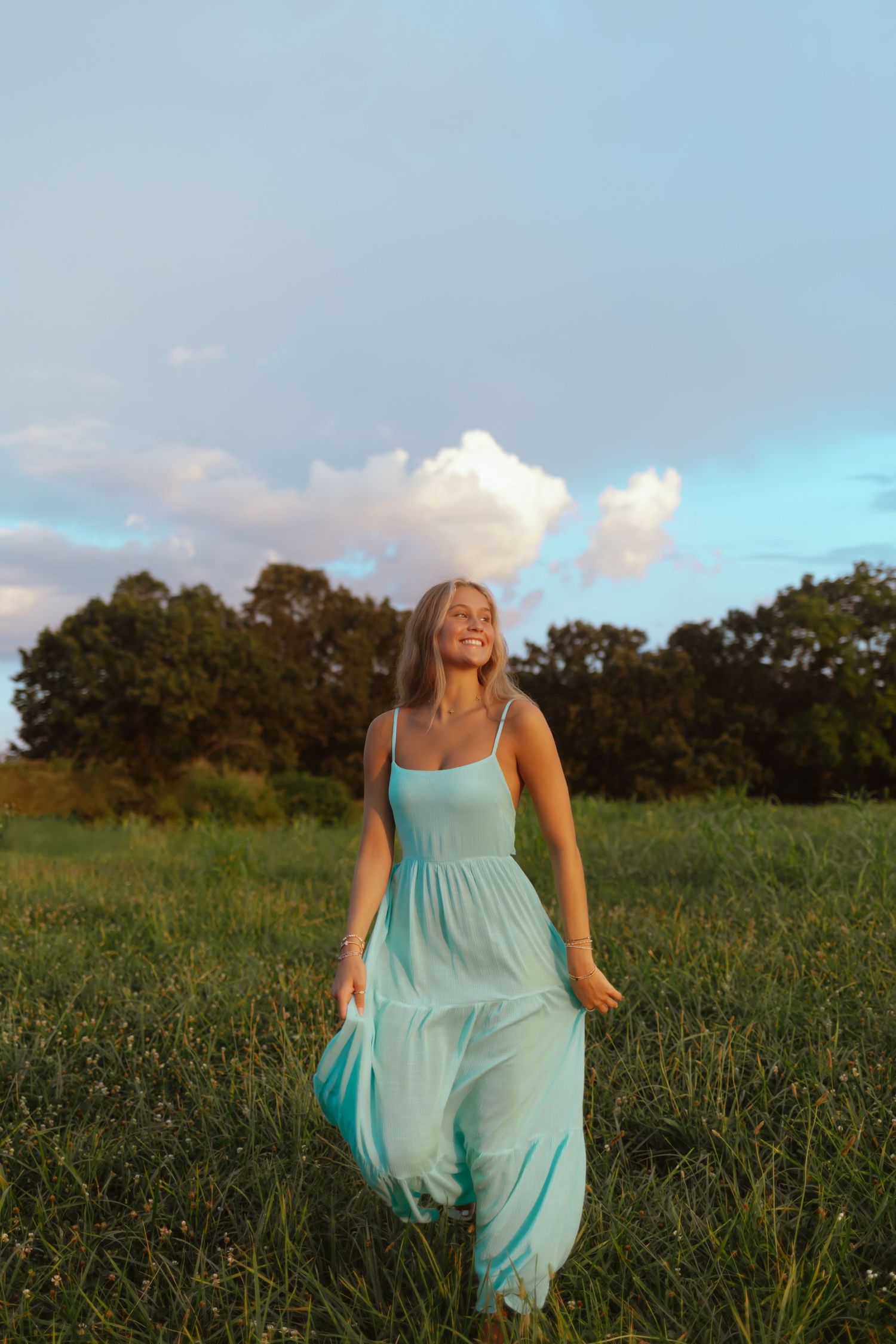 Someone wearing a flowing turquoise maxi dress stands in a grassy field at golden hour with trees in the background.