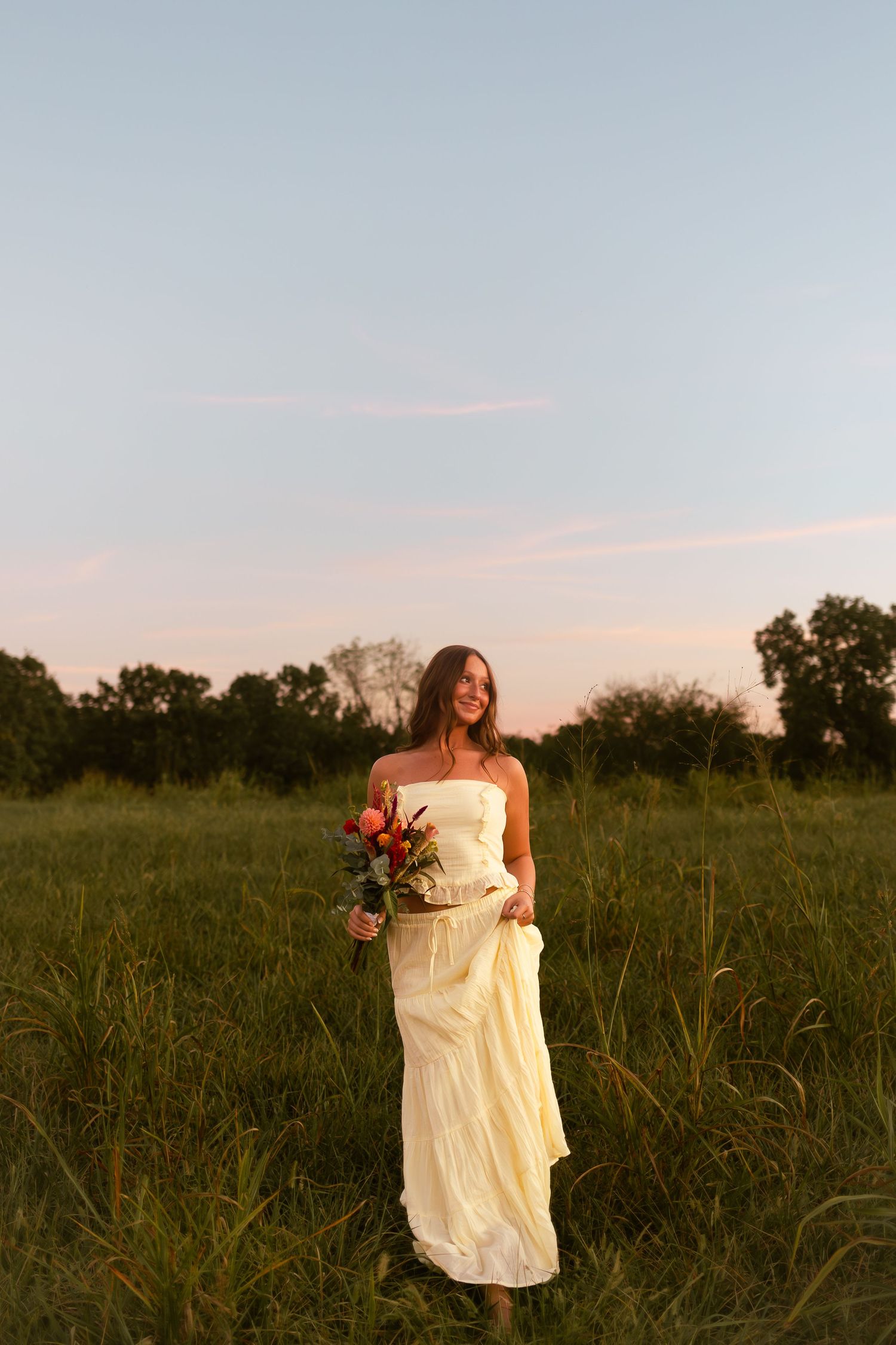 A figure in a flowing white dress holds a bouquet while standing in a sunlit meadow at golden hour.