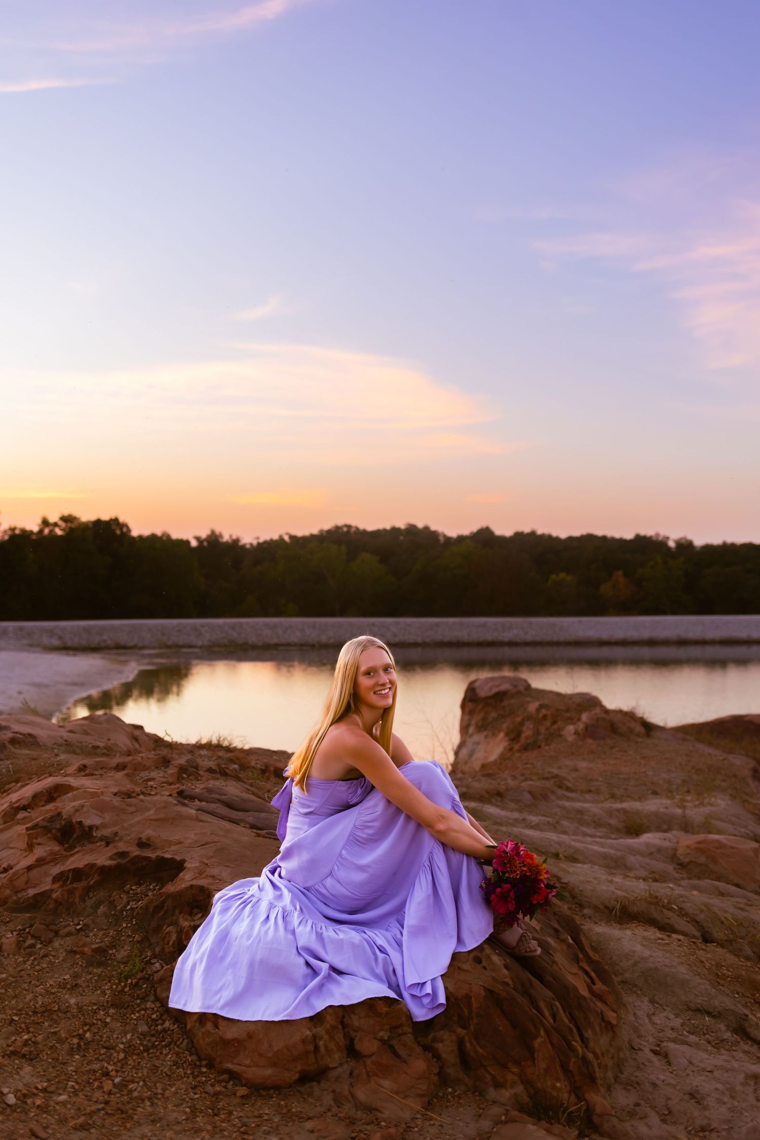 Sunset panorama showing a person in flowing purple dress posing gracefully on rocky lakeside cliffs during golden hour.