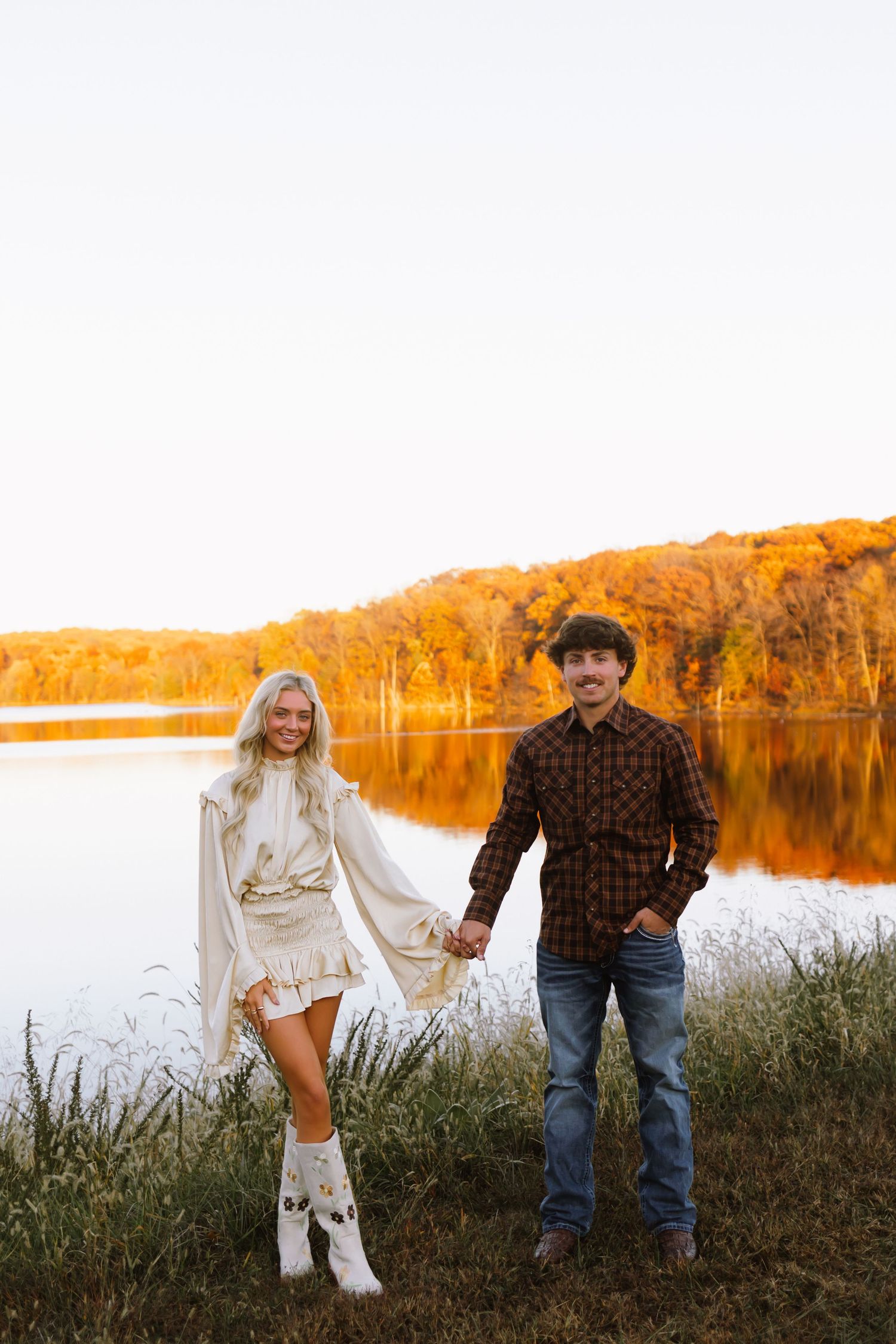 A couple walks along a lakeside path during golden hour with autumn foliage reflecting in the calm water.