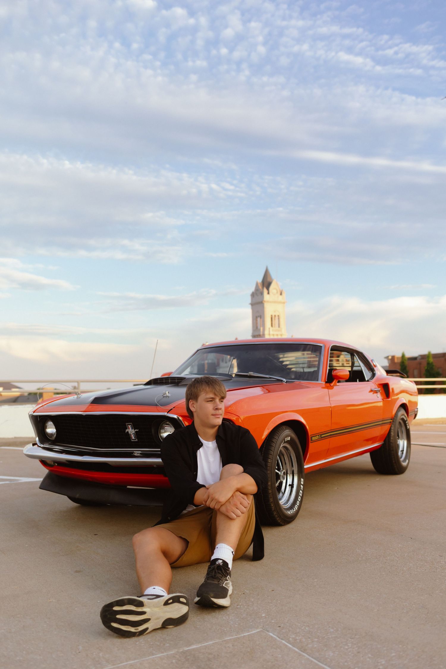 Vintage red Ford Mustang parked outdoors against cloudy sky with tower visible in background.