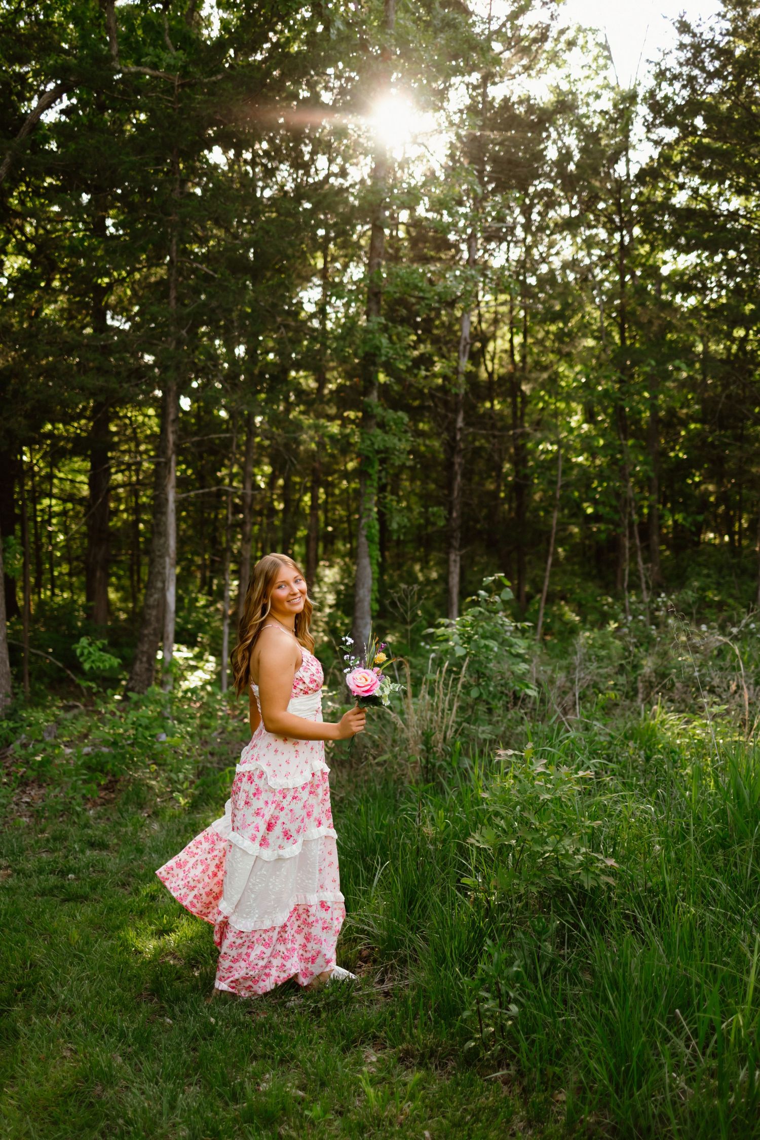 Young woman in floral pink dress holding roses, standing in sunlit forest clearing with lush green trees.