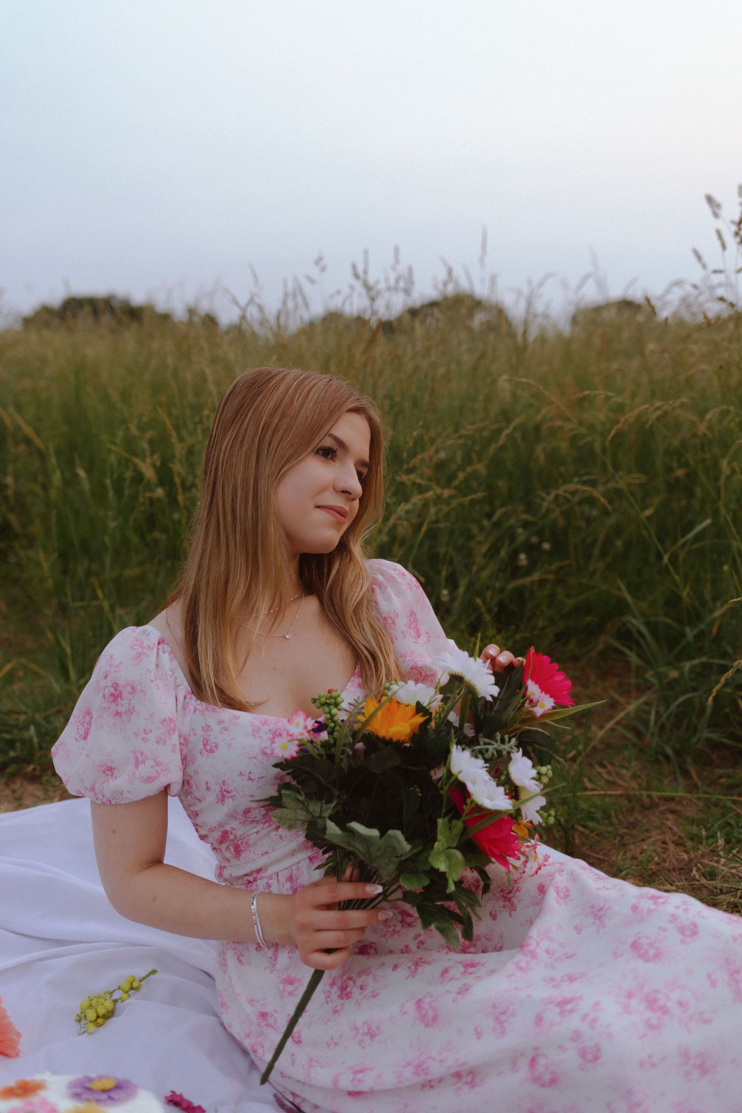 A person in a pink floral dress sits on a white blanket in a grassy field holding a colorful bouquet of wildflowers.