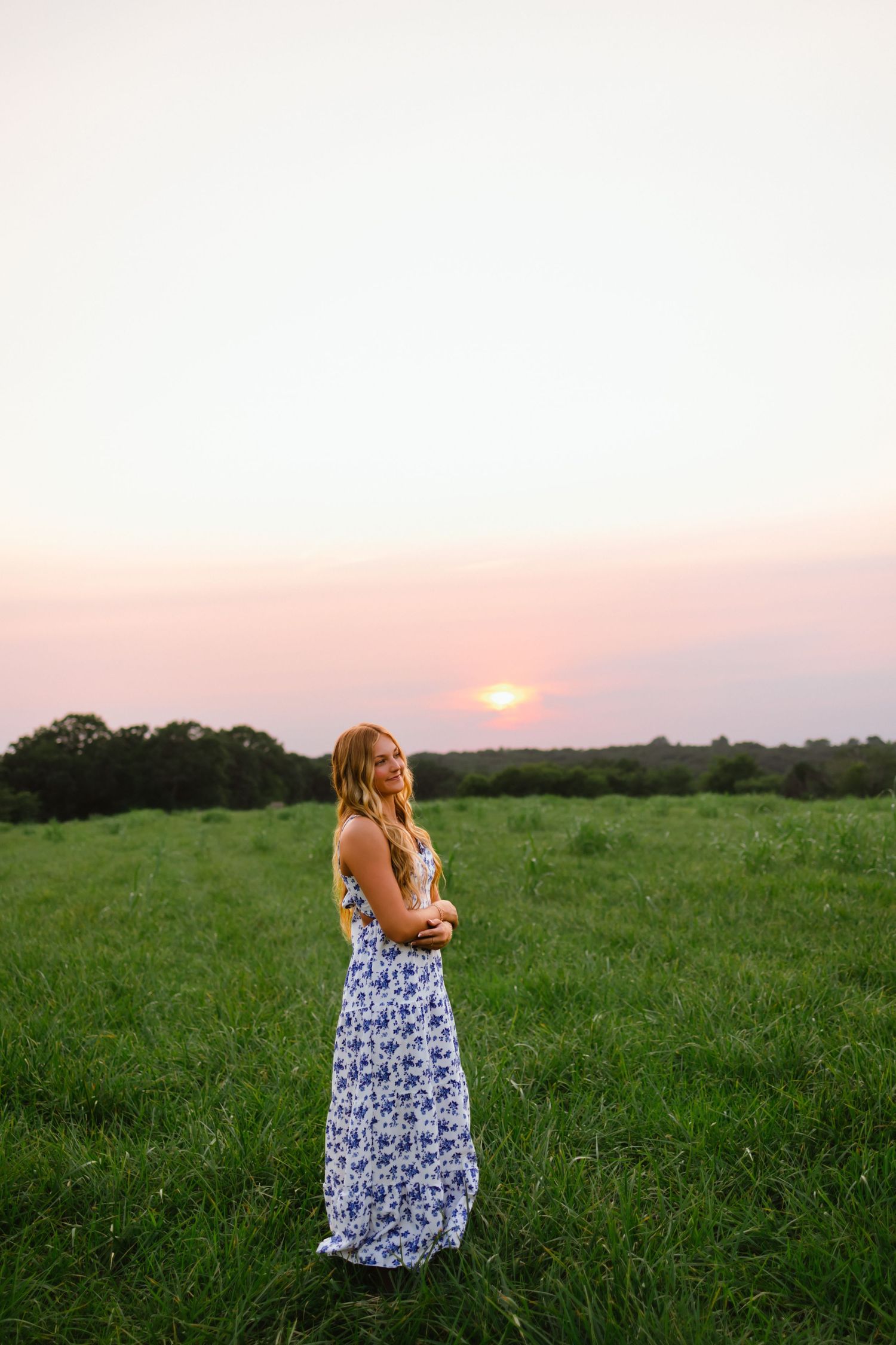 A young woman in a floral maxi dress stands in a green field at sunset.