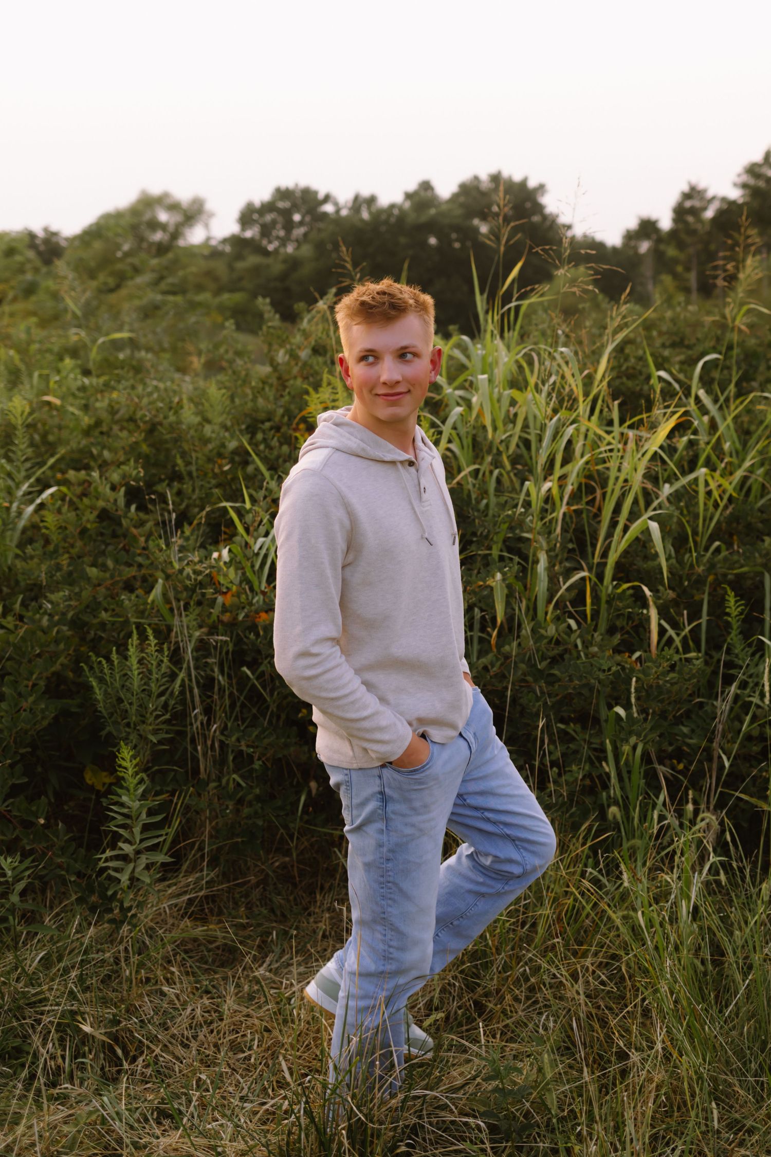 A young child in a gray sweatshirt and light jeans sits in tall grass and wildflowers at golden hour.