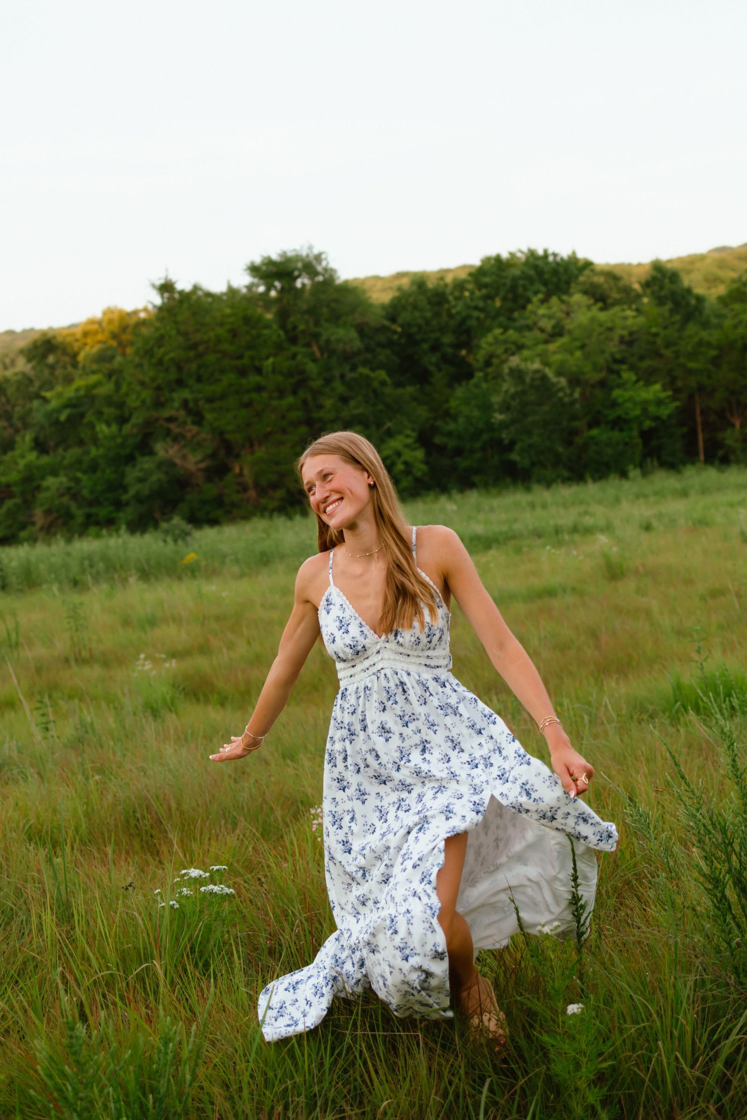 Smiling blonde woman in floral maxi dress dancing in green meadow with trees and hills in background.