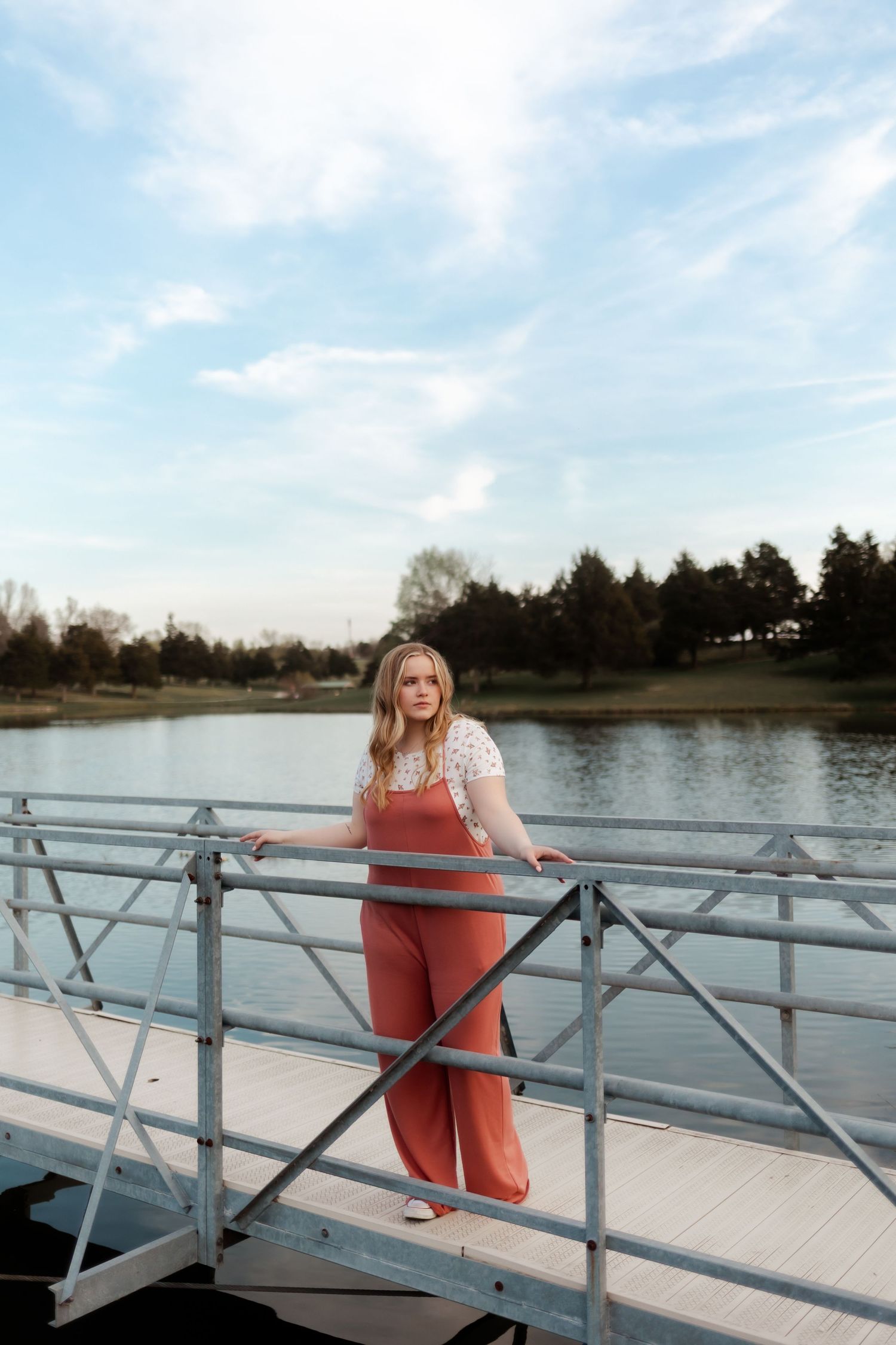 A person in coral-colored clothing sits on a white boat dock overlooking a serene lake on a sunny day.