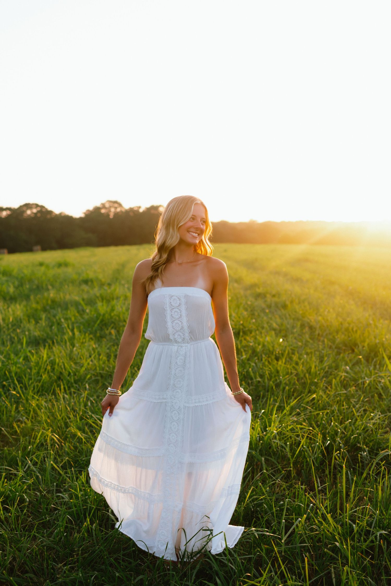 Woman in white strapless maxi dress standing in green field during golden hour sunset.