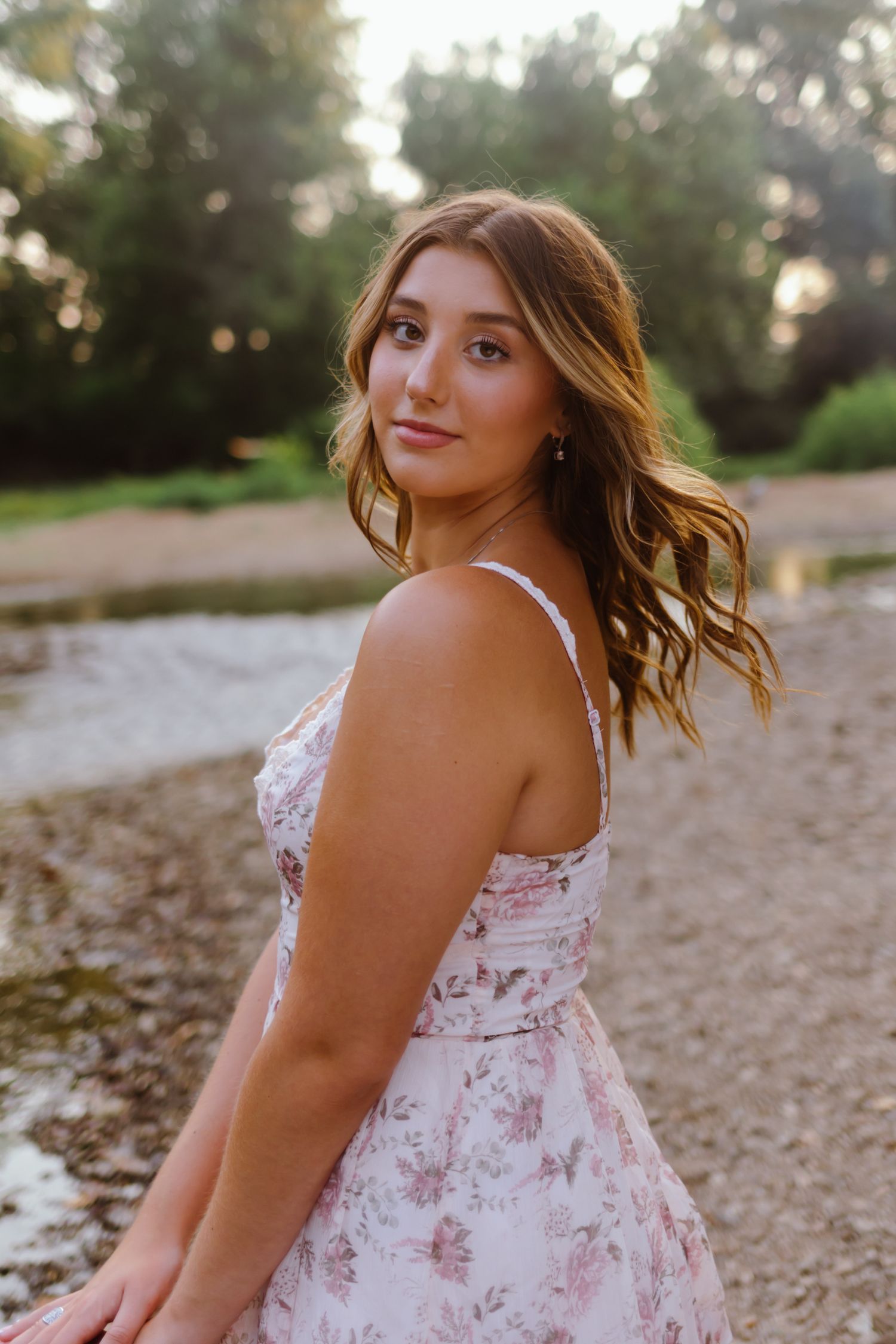 Someone in a floral summer dress poses outdoors on a rocky beach at sunset in romantic lighting.