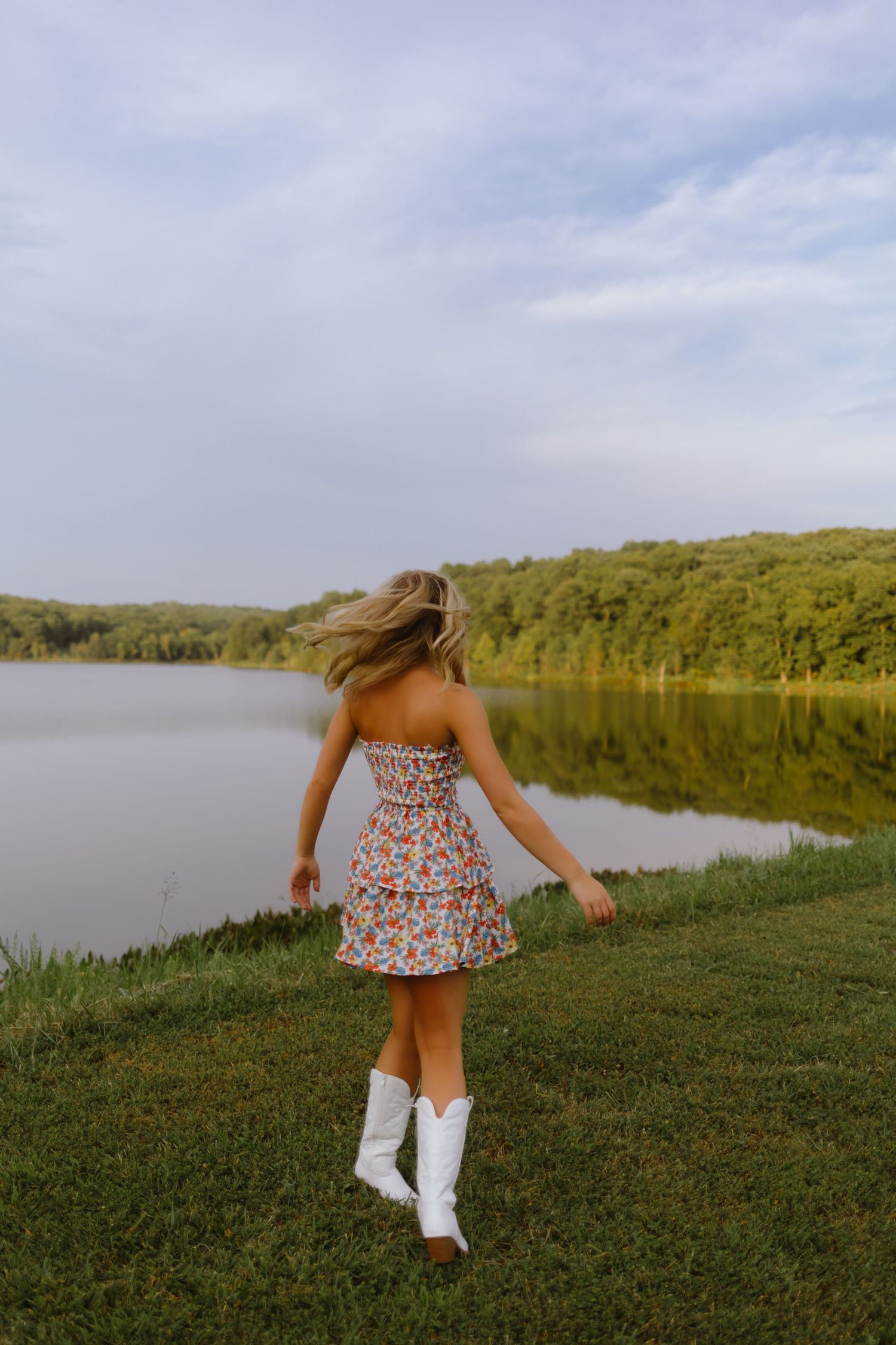 Person in floral dress and white boots spins by a serene lake surrounded by green hills on a summer evening.