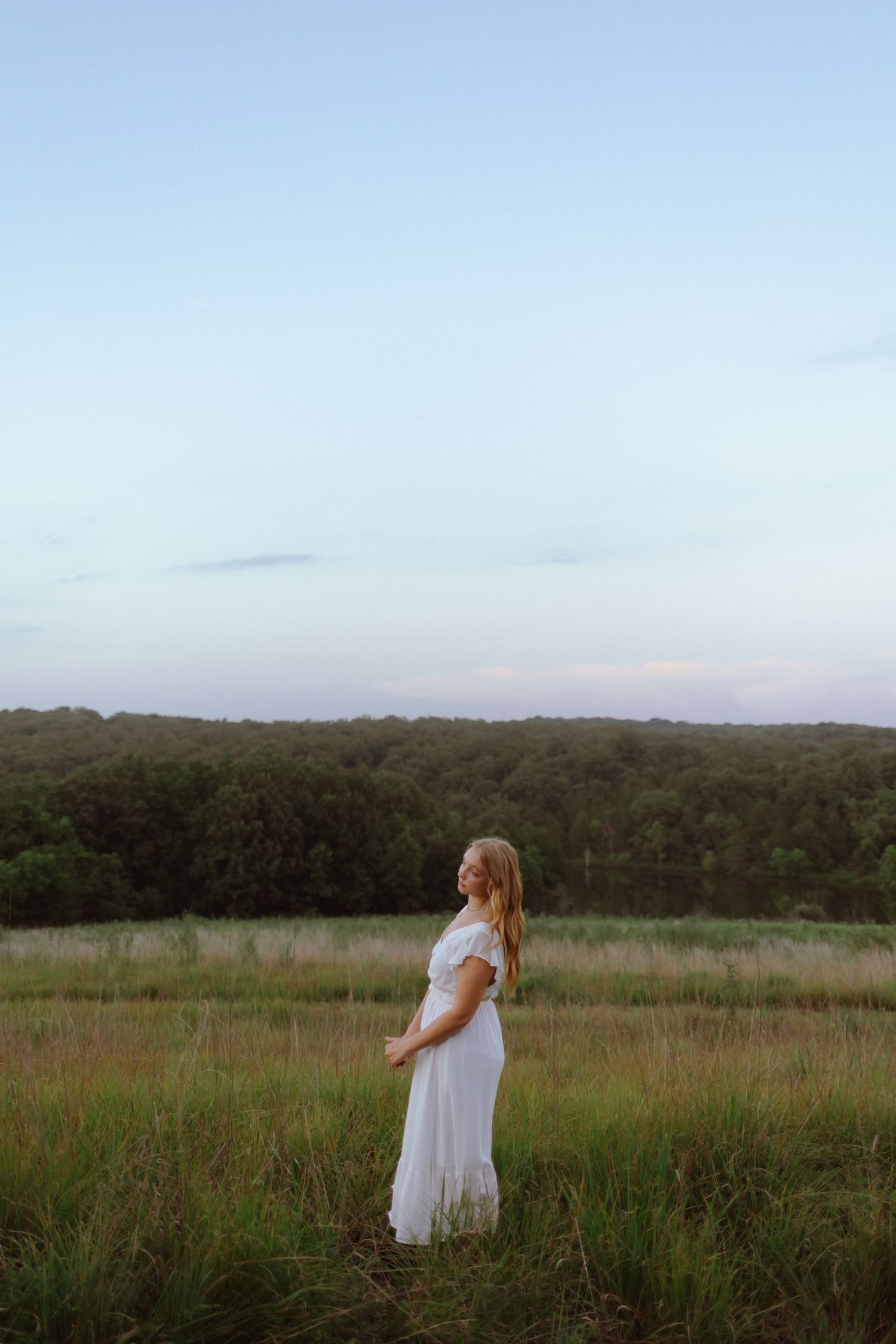 A figure in a flowing white dress stands in a serene meadow with rolling hills and forest in the background at dusk.