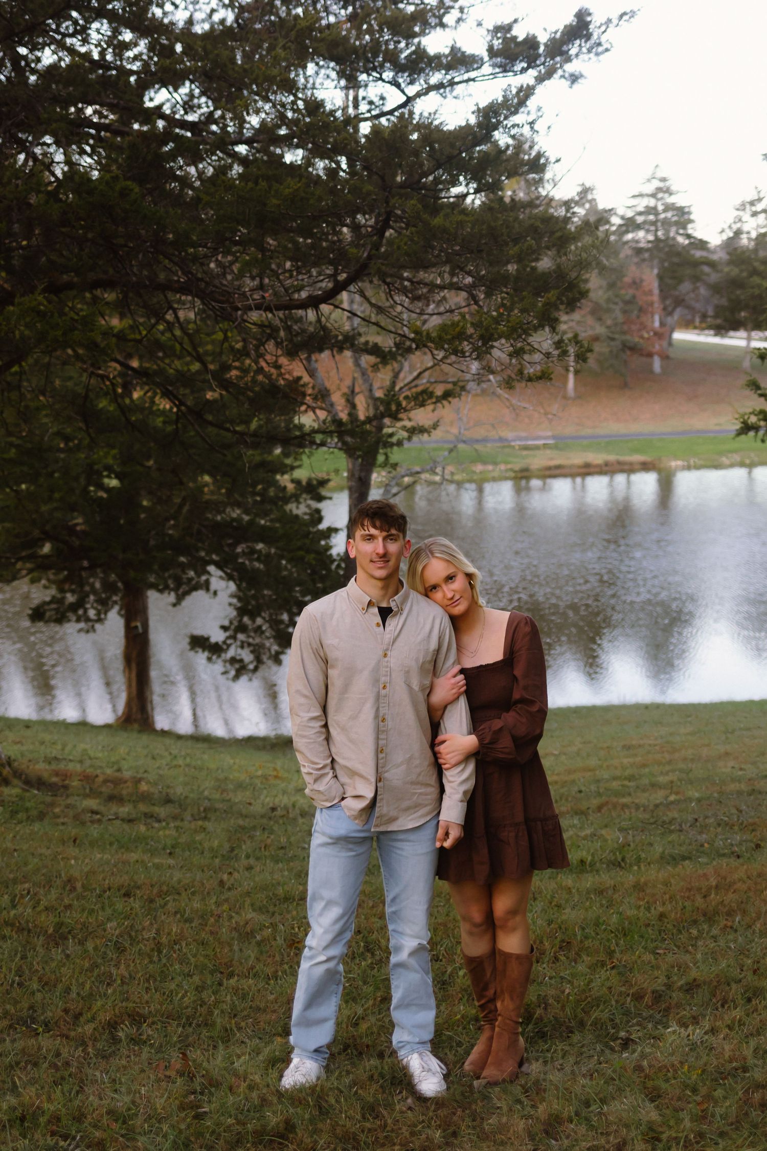 A couple stands together on a grassy hill overlooking a serene lake with pine trees in the background during autumn.