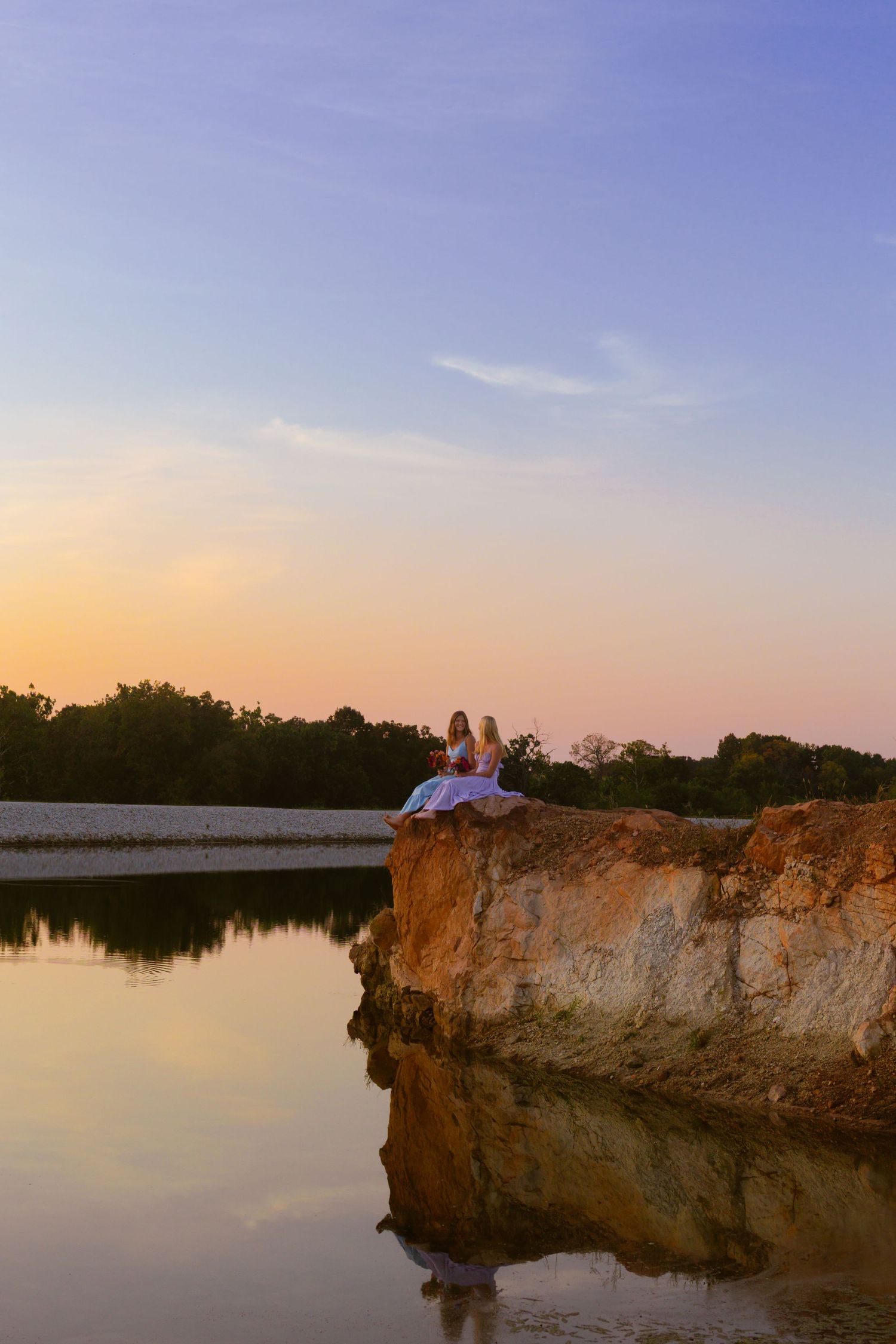 Silhouettes of people sitting on limestone cliffs overlooking a peaceful lake at sunset with warm orange sky.