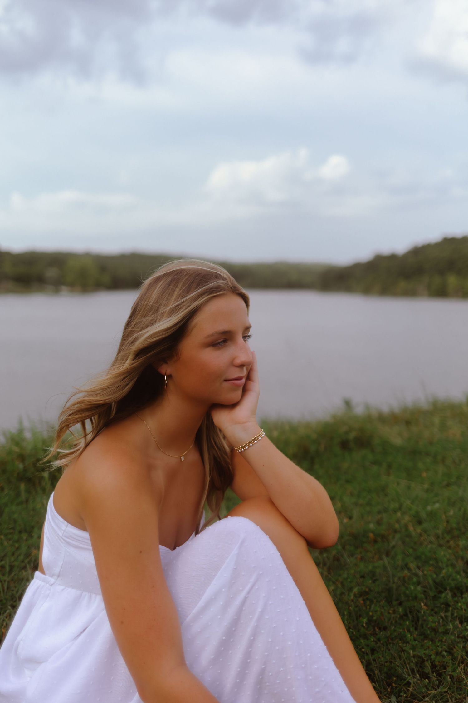 A person in a white dress sits thoughtfully on grass near a serene lake under an overcast sky.