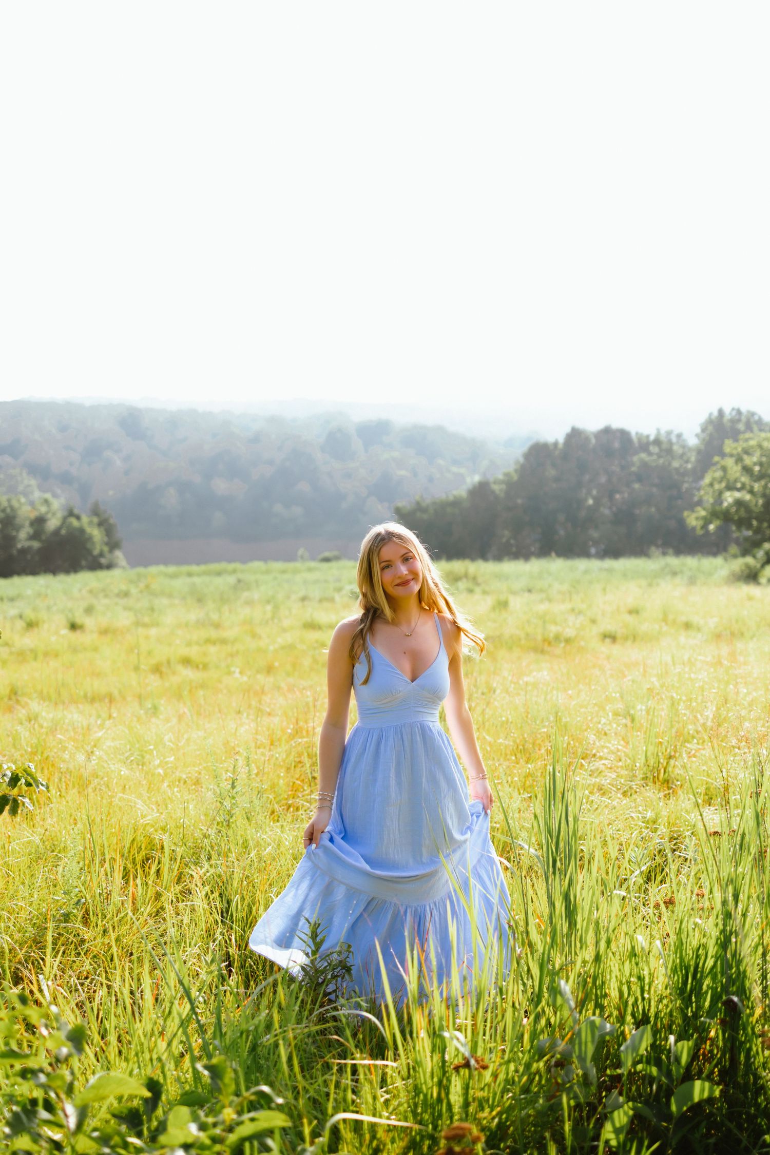 Young blonde woman in a flowing blue sundress standing in a sunny green meadow with trees in the background.