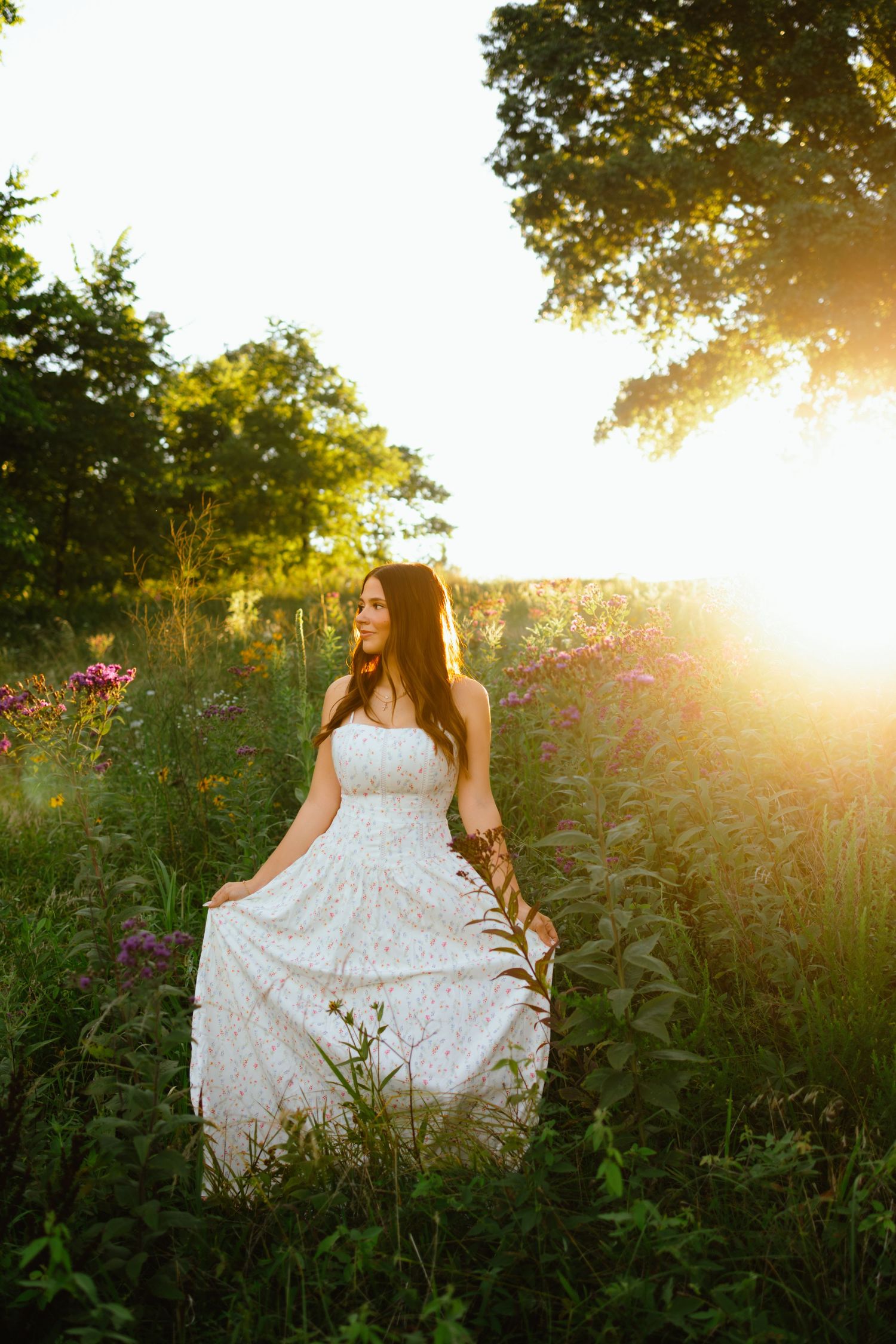 A bride in a white lace gown stands in a sunlit wildflower field at golden hour.