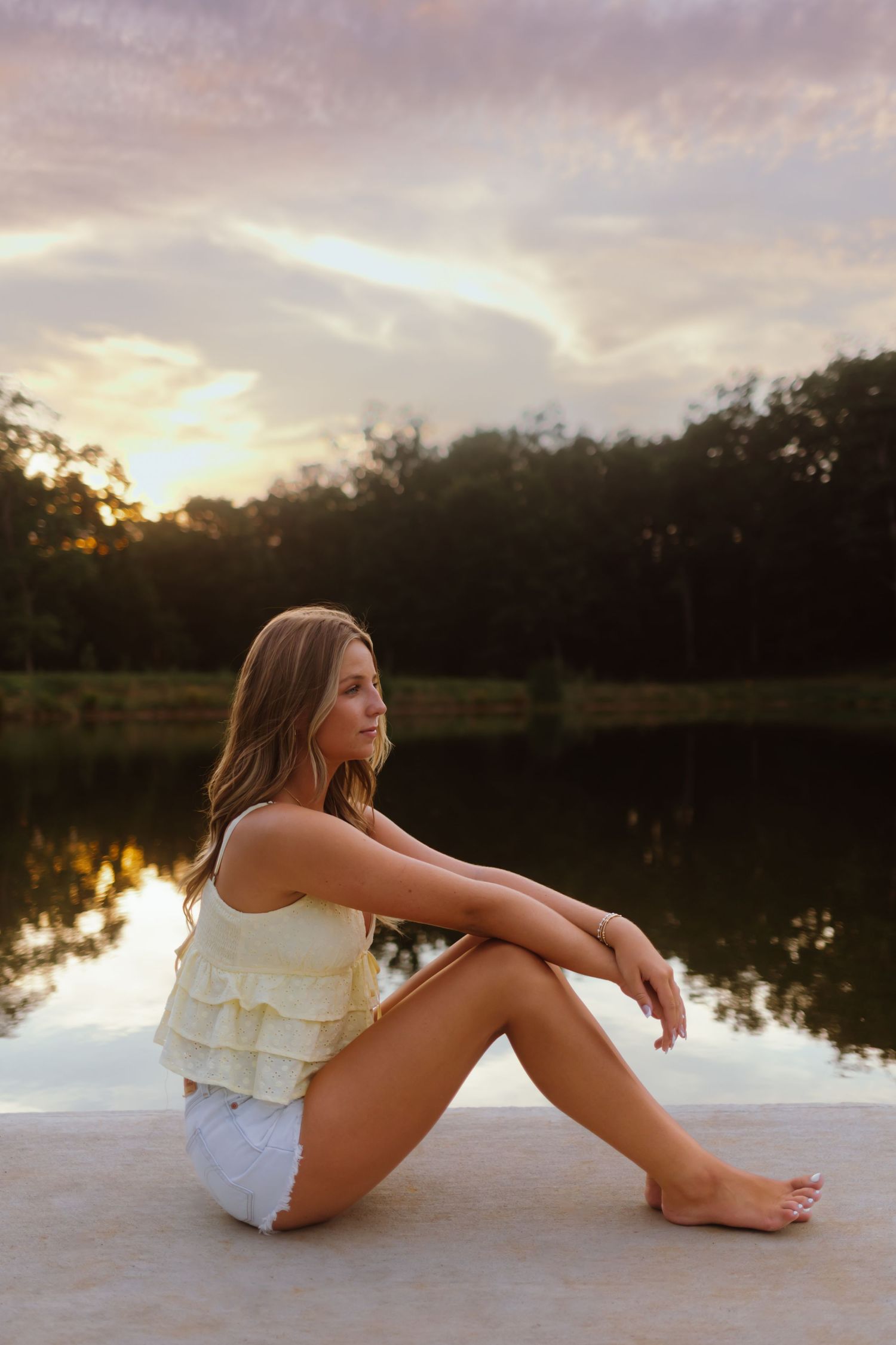A person in white summer clothing sits on a dock by a calm lake at sunset with trees reflected in the water.