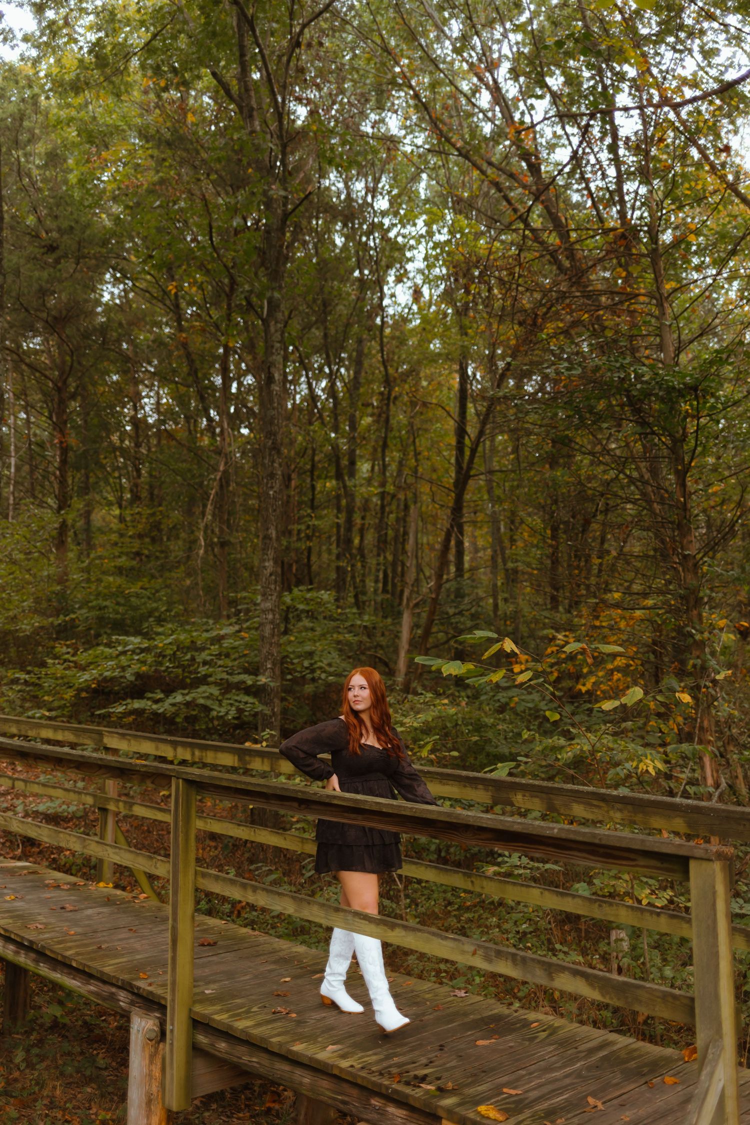 A figure in a black dress and white boots stands on a wooden boardwalk surrounded by autumn forest scenery.