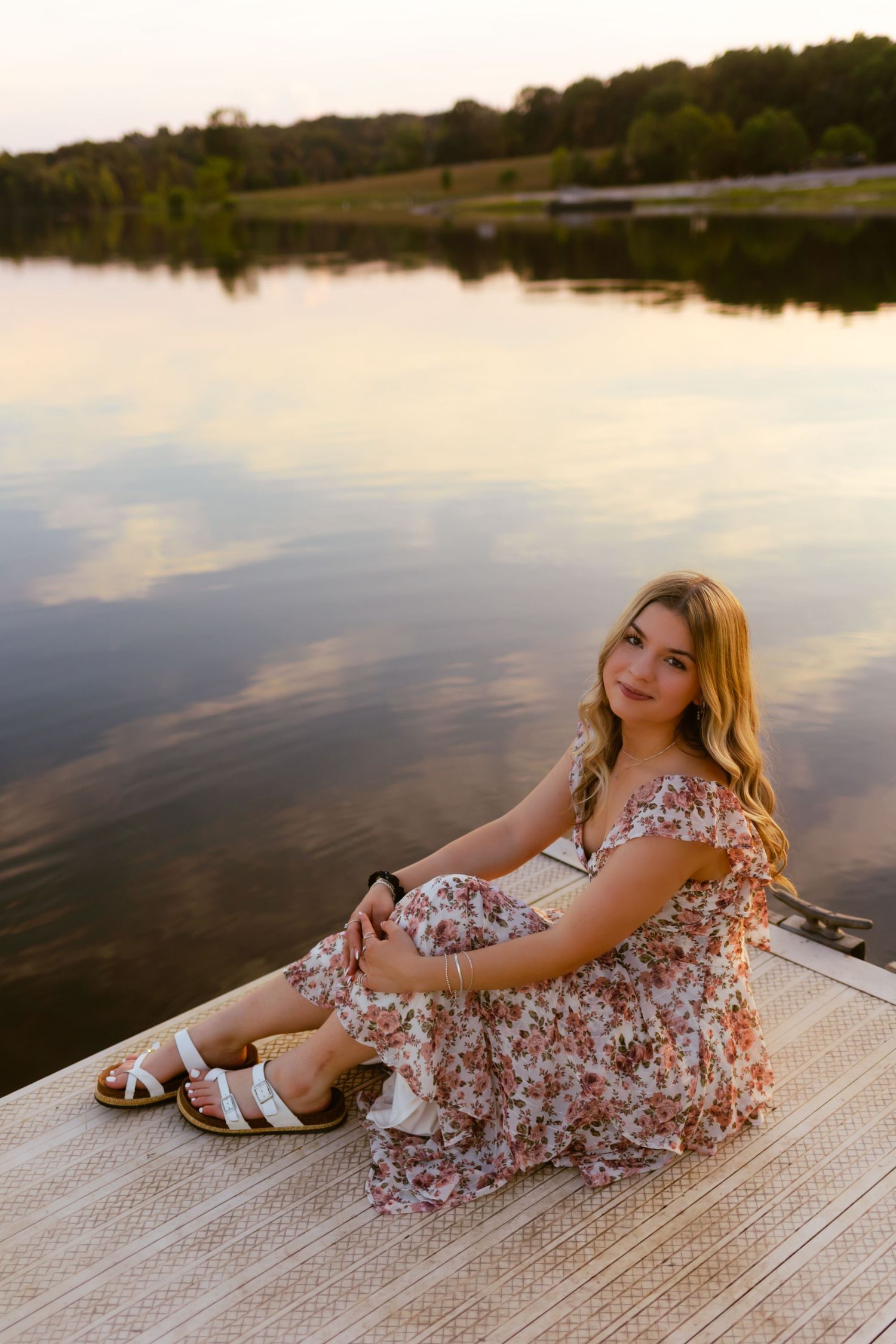 A person in a floral dress sits on a dock by a serene lake at sunset with reflective water in the background.