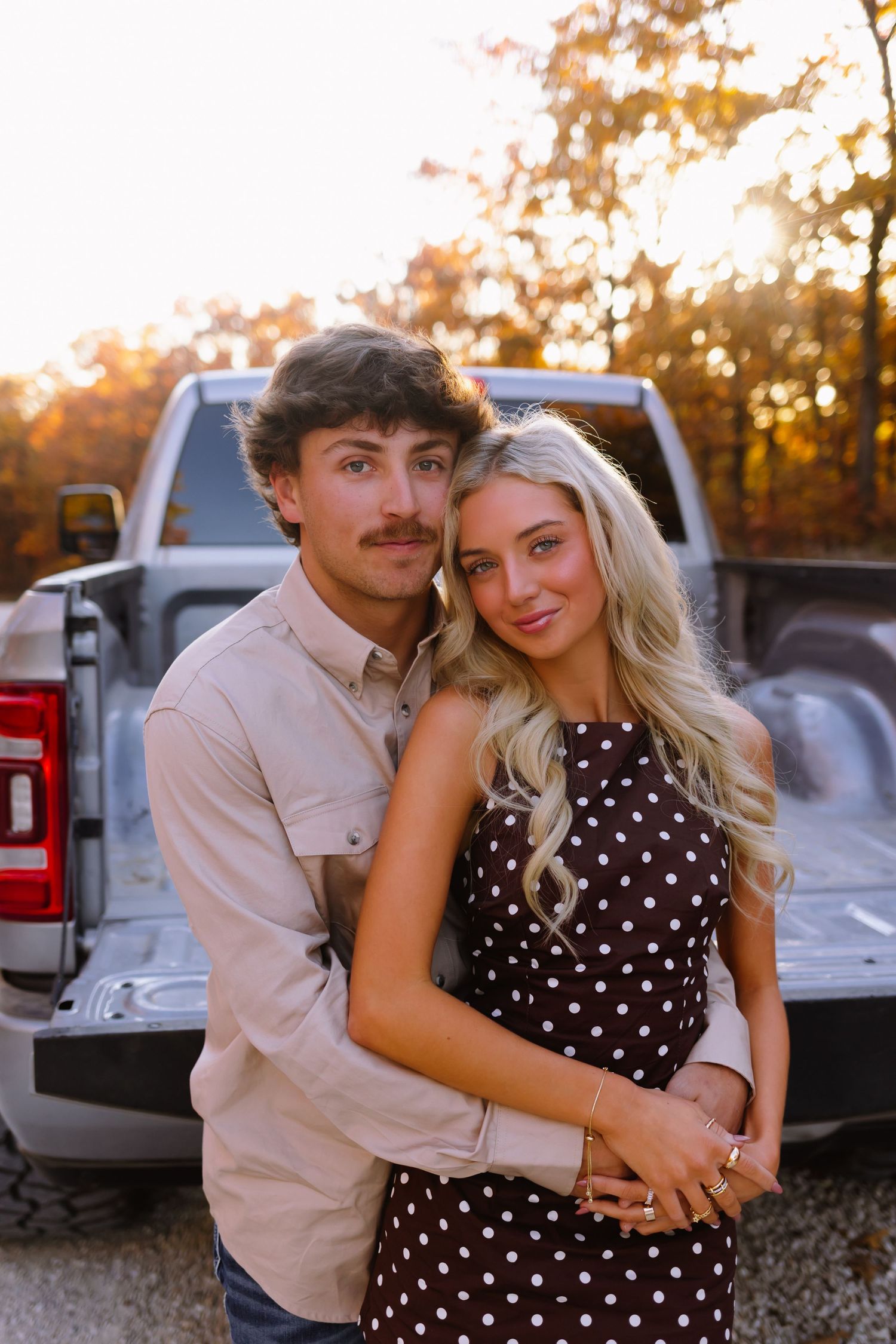 A couple poses together by a pickup truck during golden hour sunset in a polka dot dress and casual attire.