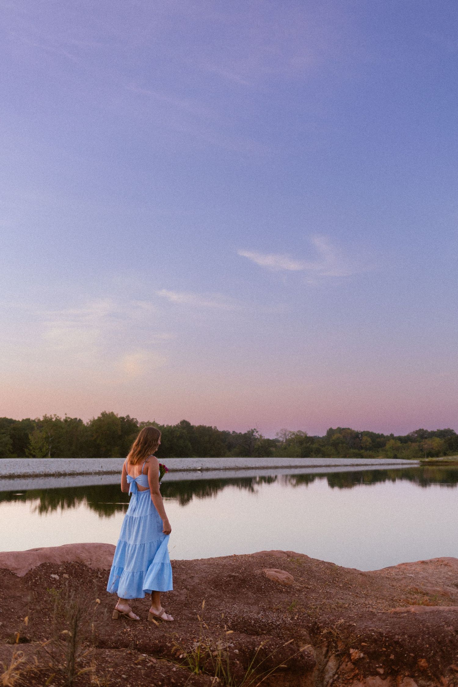 A woman in a blue sundress stands on the shore of a serene lake during sunset with trees in the background.
