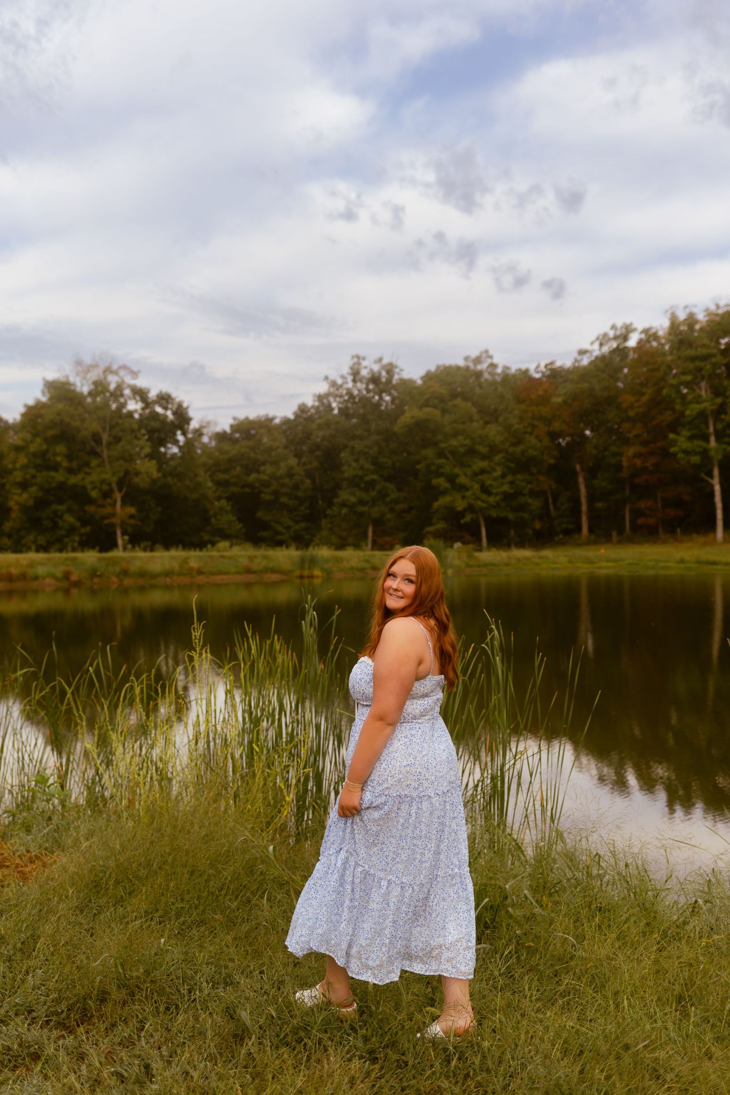 A person in a light blue sundress stands beside a serene pond surrounded by trees on a summer day.