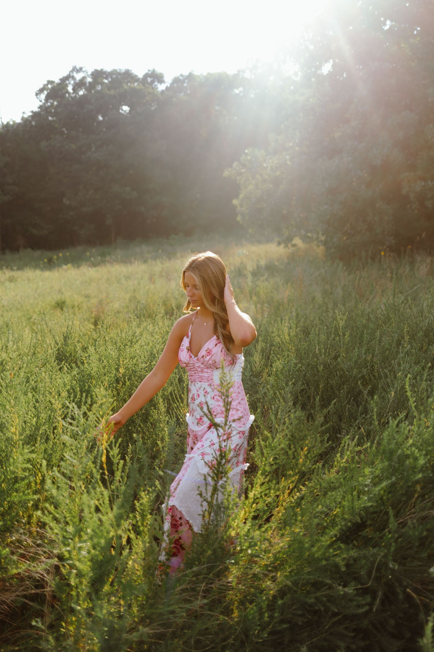 A person in a floral summer dress walks through a sunlit meadow of tall wildflowers and grass during golden hour.