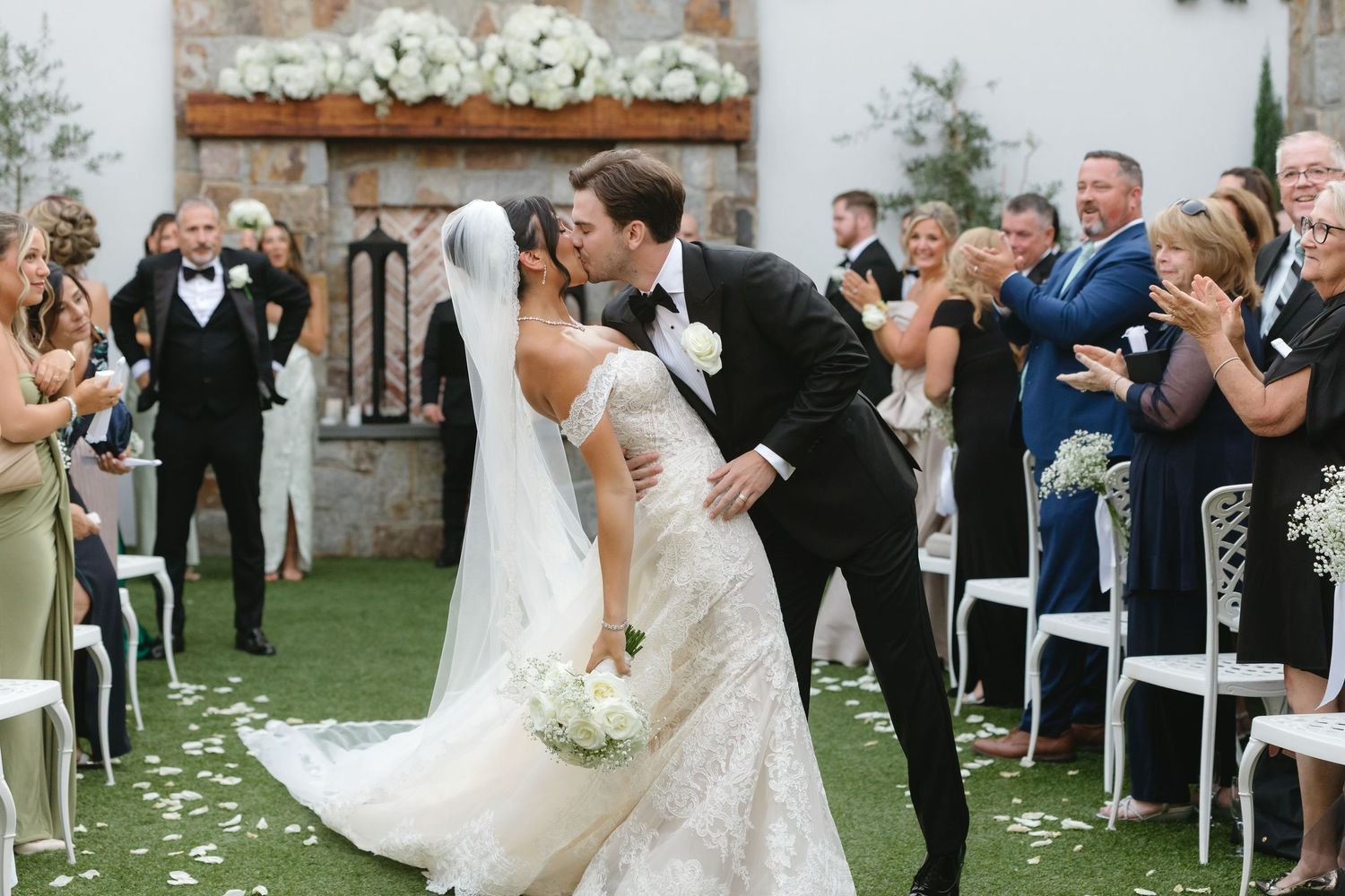 Wedding ceremony guests seated in white chairs on a lawn with flower petals scattered along the aisle.