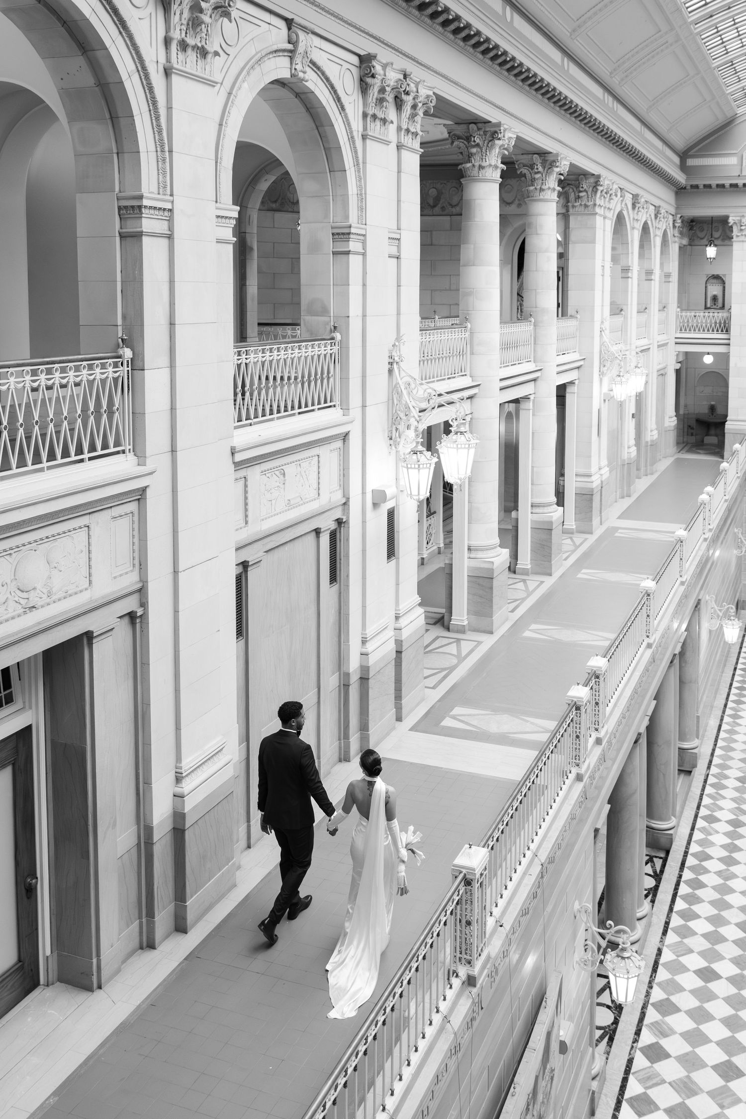 A black and white architectural interior view shows a long corridor with checkered floors and arched columns along the walls.