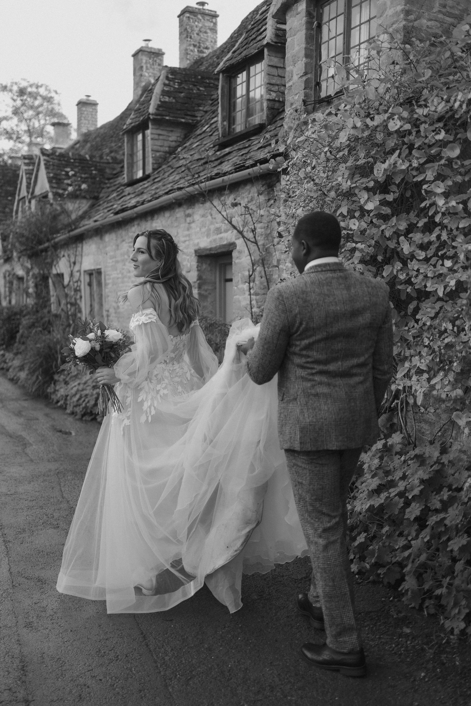 A couple in wedding attire walks along a charming stone cottage path in black and white.