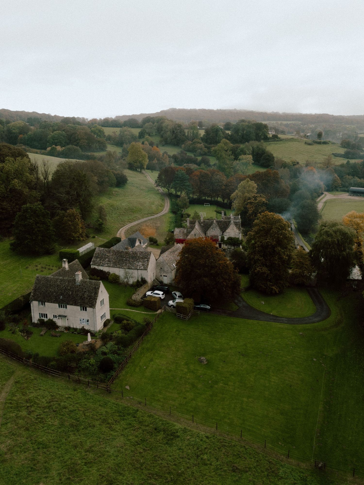 Aerial view of historic stone manor houses nestled in rolling green countryside with winding roads and autumn trees.