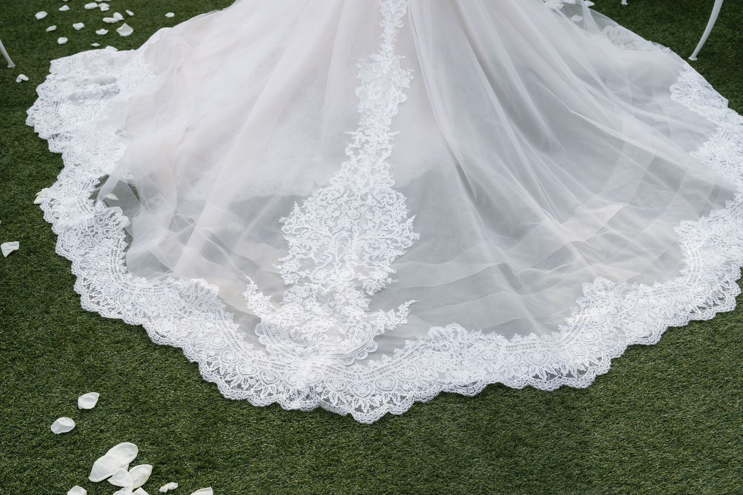 Elegant white lace wedding dress train with intricate floral embroidery details spreads across green grass.