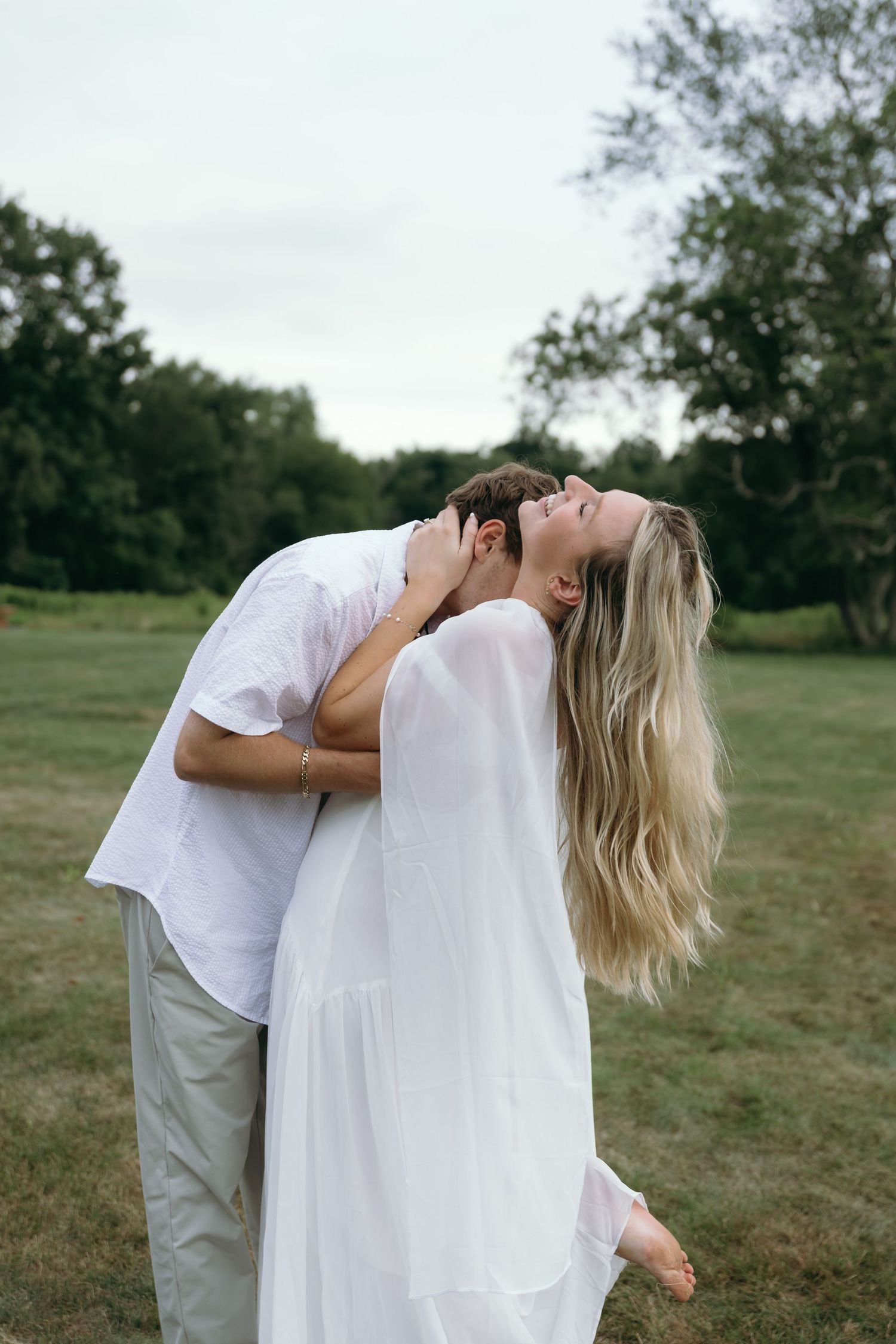 A romantic couple in white clothing share an intimate dip kiss pose in a grassy outdoor field.