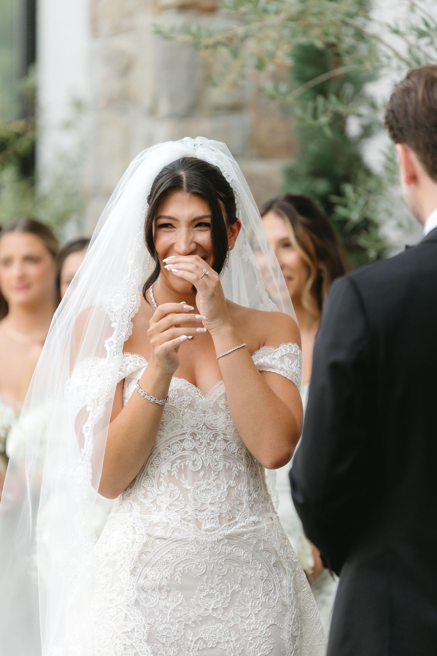 A bride in a white lace wedding dress laughs joyfully during an outdoor ceremony.