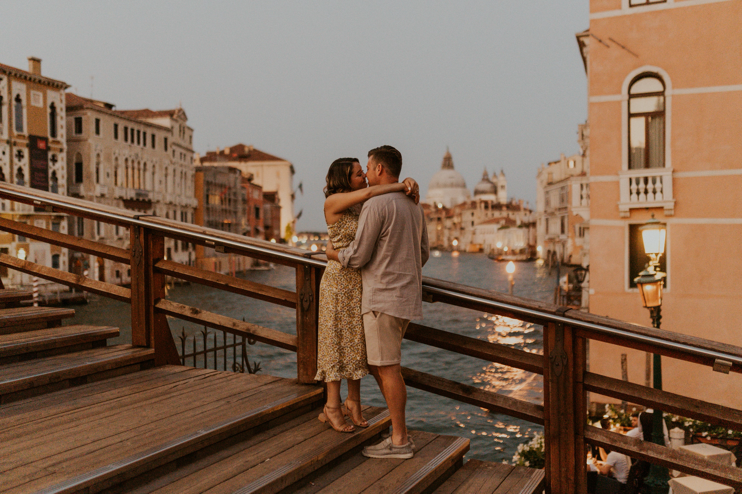 A couple stands on a wooden bridge in Venice, Italy at sunset with historic buildings in the background.