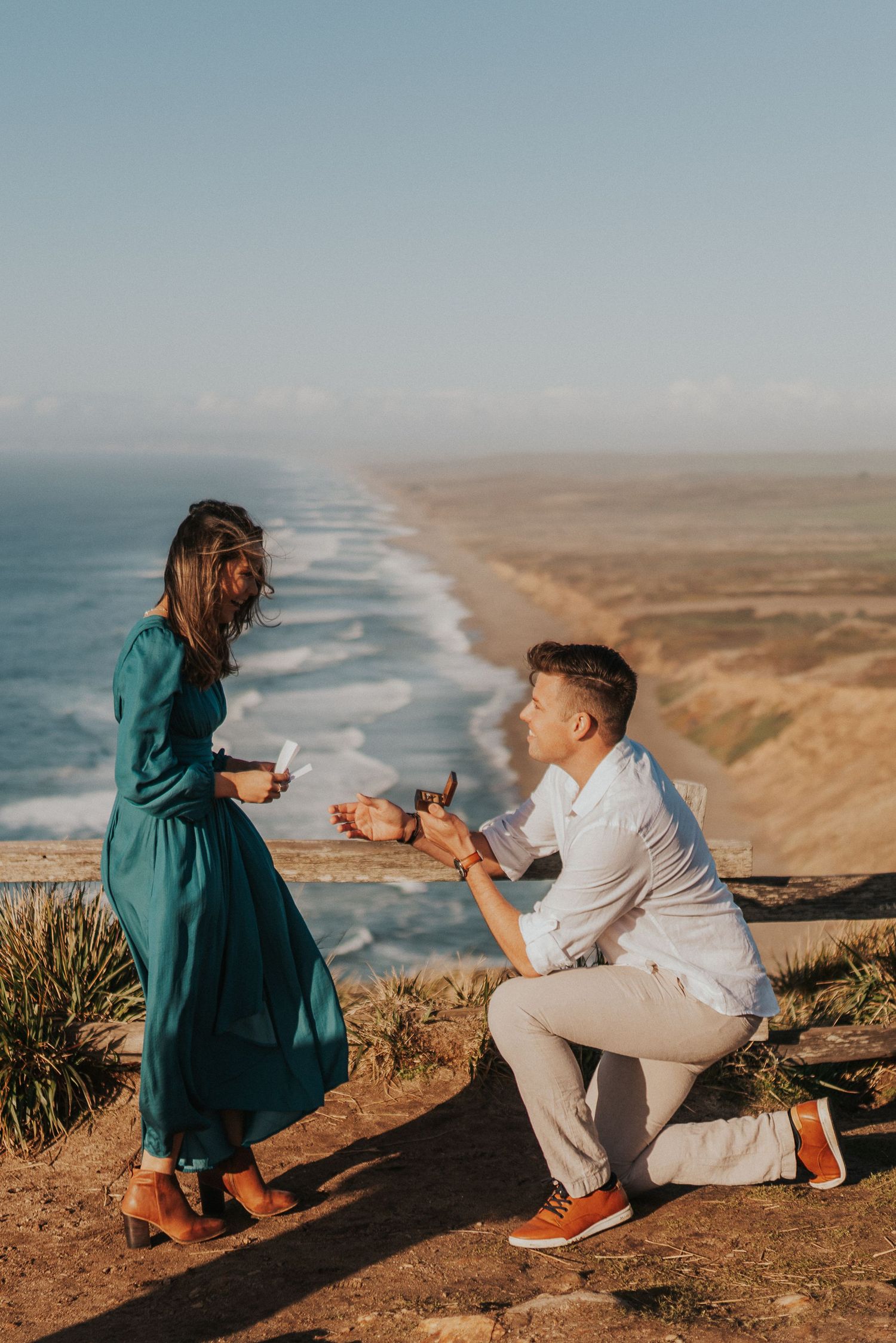 A romantic proposal scene on a coastal cliff overlooking a dramatic beach and ocean landscape at sunset.