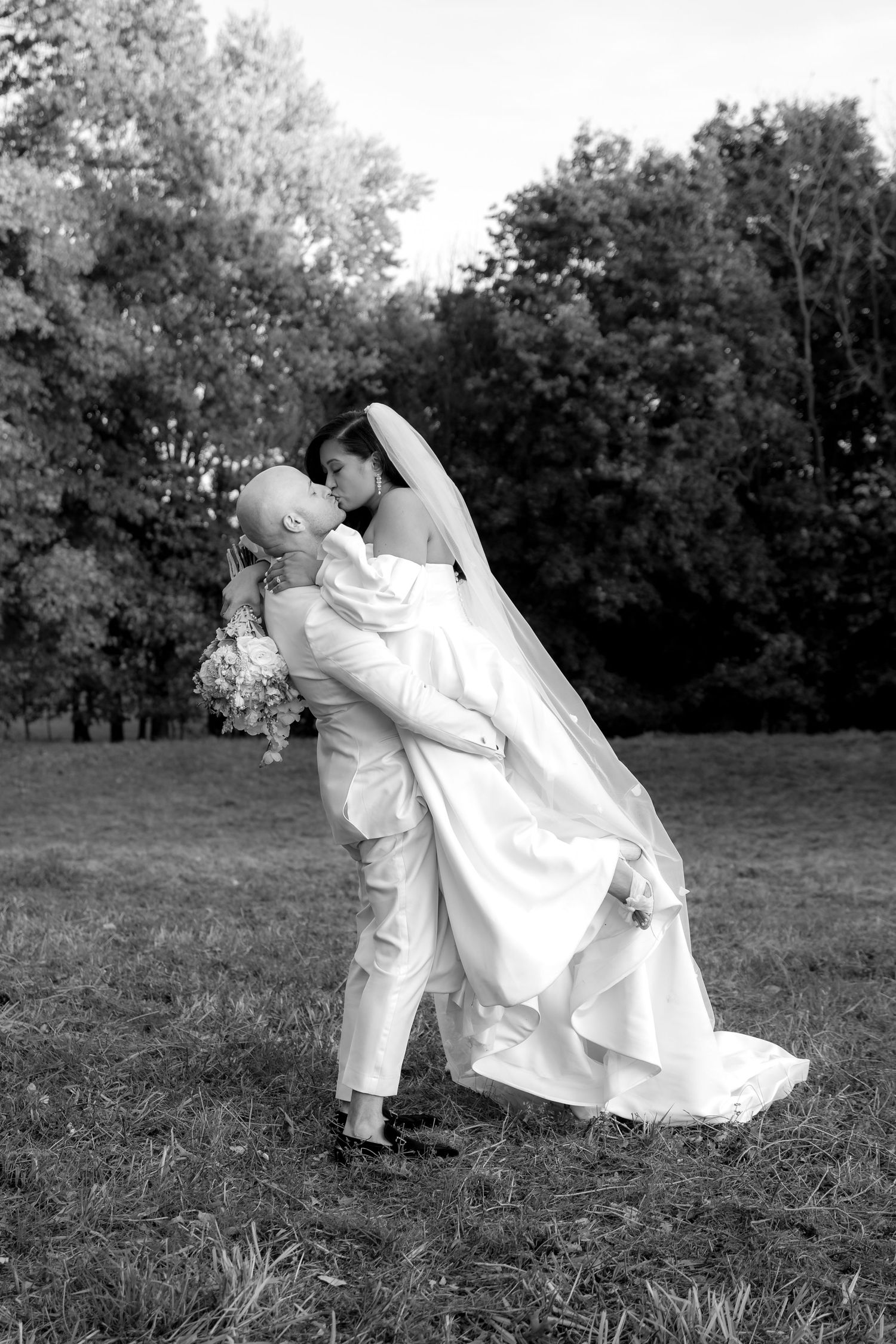 A romantic black and white wedding photo showing a couple embracing outdoors with trees in the background.