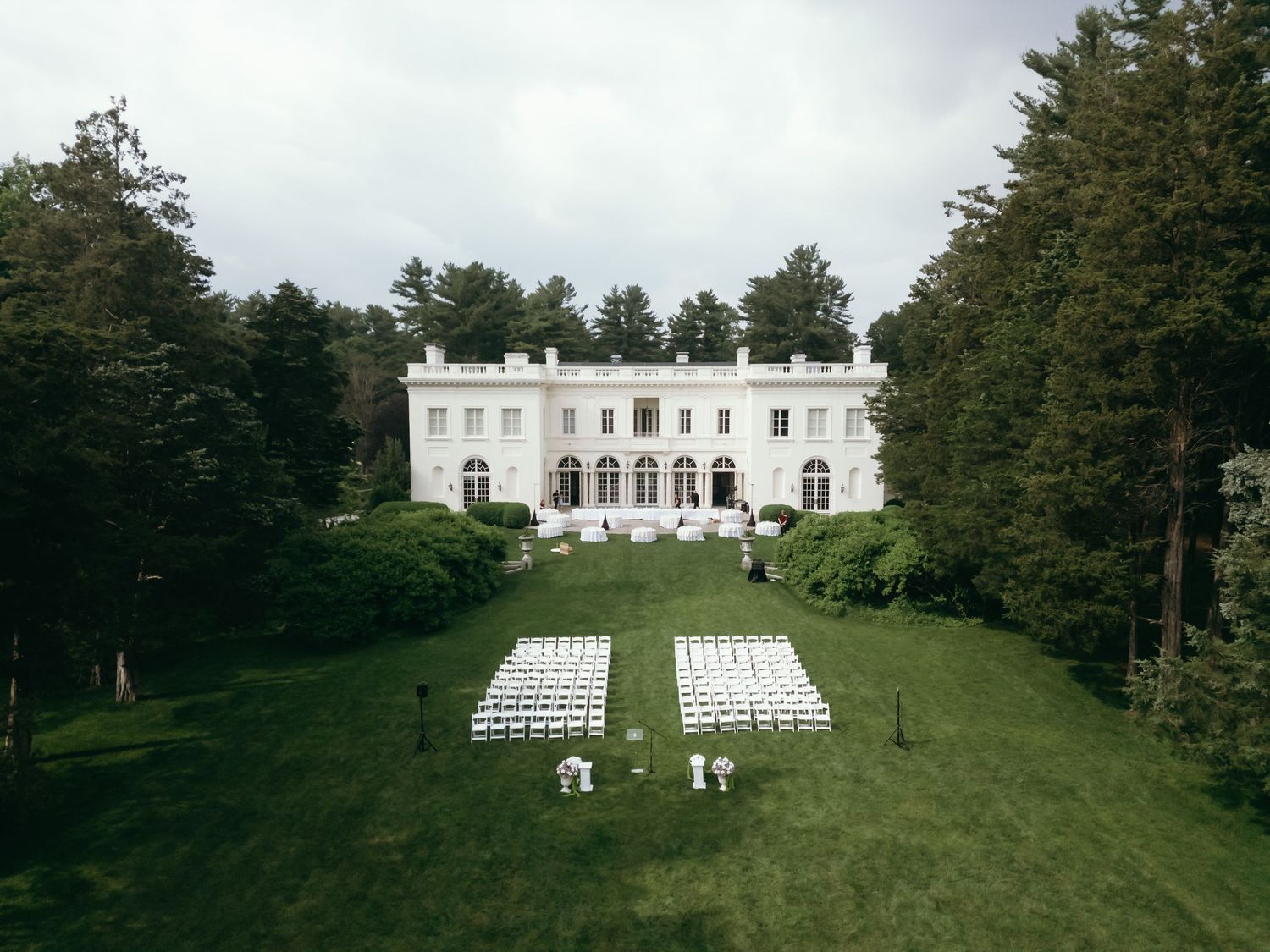White chairs arranged on a green lawn for an outdoor wedding ceremony with a stately white mansion in the background.